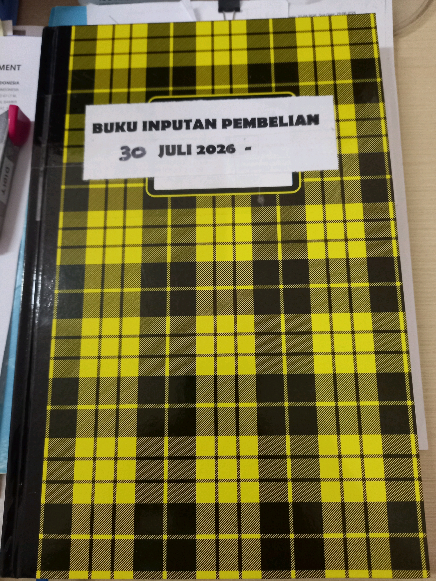 The image shows a close-up, top-down view of a notebook with a yellow and black plaid pattern. A white label is affixed to the front cover of the notebook. The label contains black text that reads "BUKU INPUTAN PEMBELIAN" in all caps, followed by "30 JULI 2026 -" on the line below. The notebook is lying on a desk. To the left of the notebook, a portion of another document with text is visible, and further left, a pink highlighter pen is partially visible. The background appears to be a wooden desk surface. There is no identifiable location in Samarinda, Indonesia.