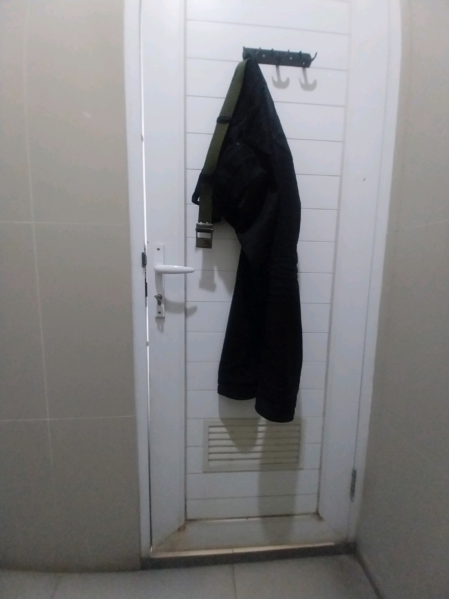A black jacket with a green strap is hanging on a wall-mounted hook on a white, paneled door. The door has a white handle and a small lock mechanism. To the left of the door, a tiled wall is visible. The floor appears to be tiled as well. The image is taken indoors, possibly in a bathroom or utility area, given the tiled surfaces and the presence of a ventilation grate below the jacket. The lighting is even, suggesting an indoor environment with artificial light. The scene is static and appears to be a casual arrangement of clothing.