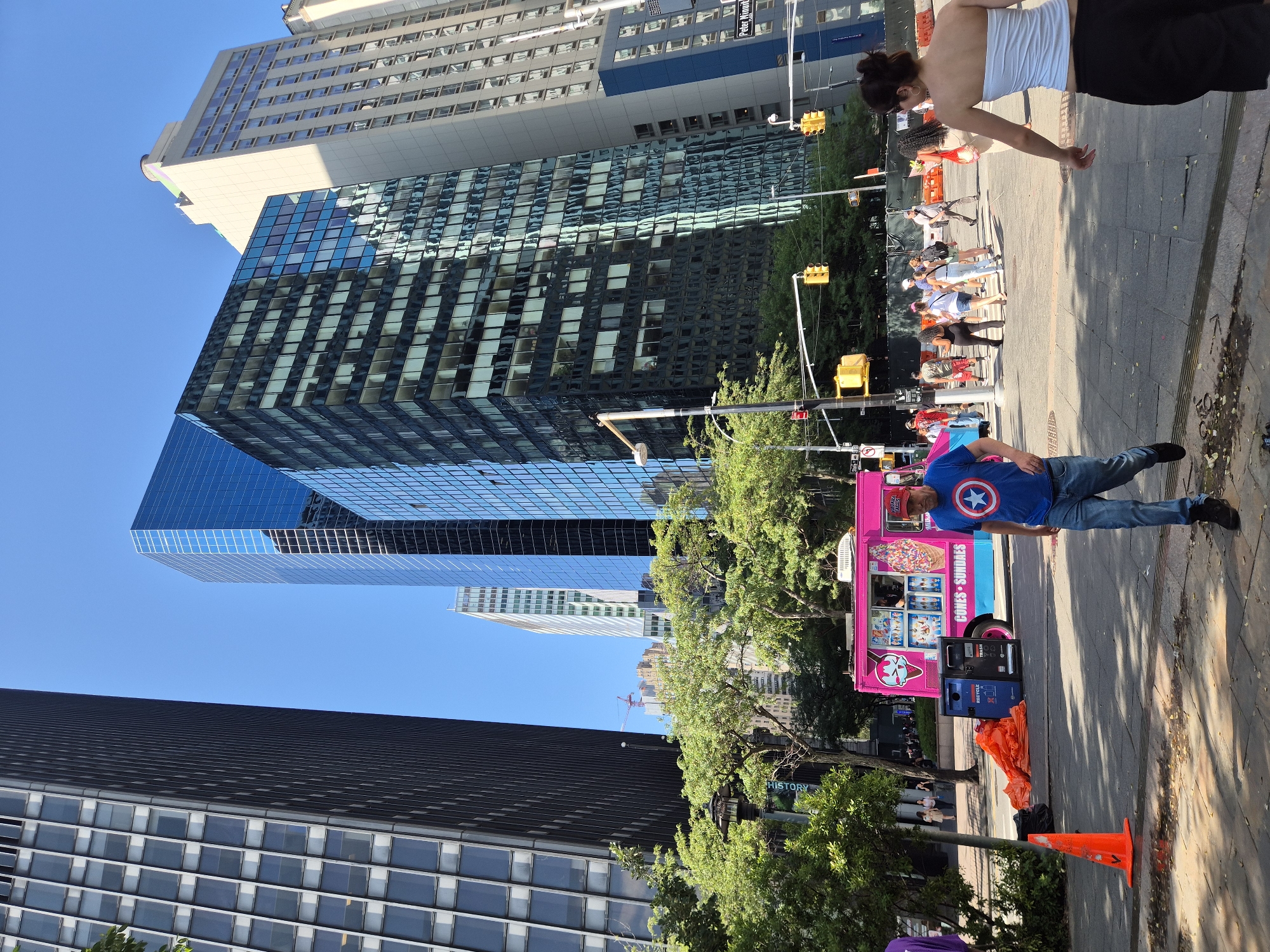 In a bustling New York City street scene, skyscrapers dominate the background under a clear blue sky. A vibrant pink ice cream truck, adorned with images of cones and sundaes, is parked on the right side of the frame. In the foreground, a man wearing a blue t-shirt with a Captain America shield logo and blue jeans walks to the right, while a woman with dark hair, wearing a white strapless top and black pants, walks to the left. Pedestrians can be seen crossing the street in the mid-ground. Orange traffic cones and construction materials are present on the sidewalk. The image captures a lively urban environment with a mix of architecture, people, and a food truck.