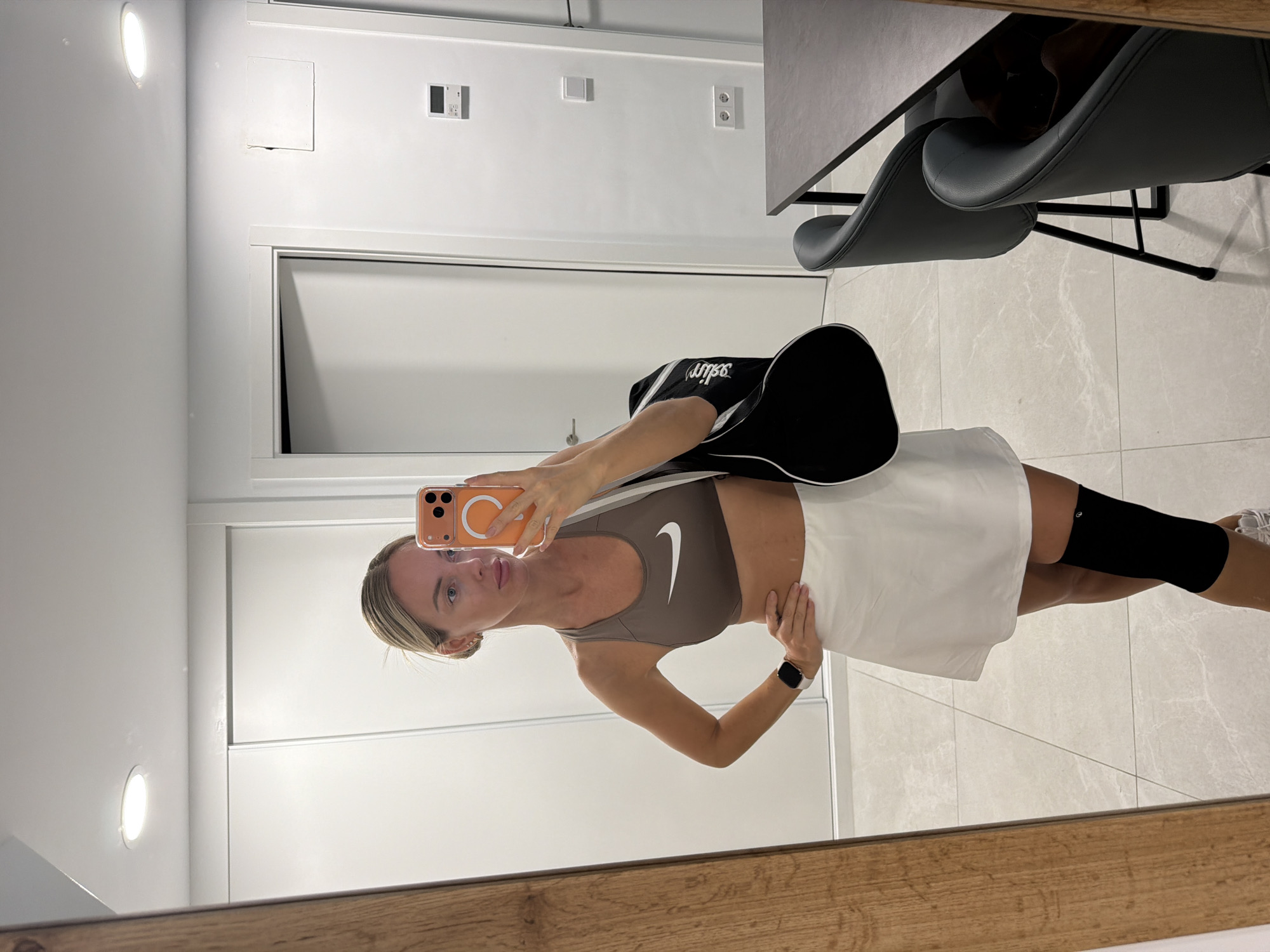 In Finestrat, Spain, a woman stands in what appears to be a modern apartment, taking a mirror selfie. She is dressed in athletic wear, including a brown Nike sports bra, a white tennis skirt, and a black knee brace. A black duffel bag is slung over her left shoulder, and she holds an orange-cased smartphone in her right hand. The background reveals a clean, white-tiled floor, a large mirror reflecting her image, a closet with sliding doors, and a doorway leading to another room. Further in the background, a dark dining table with chairs is visible, suggesting a communal living space. The lighting is bright, likely from overhead fixtures.