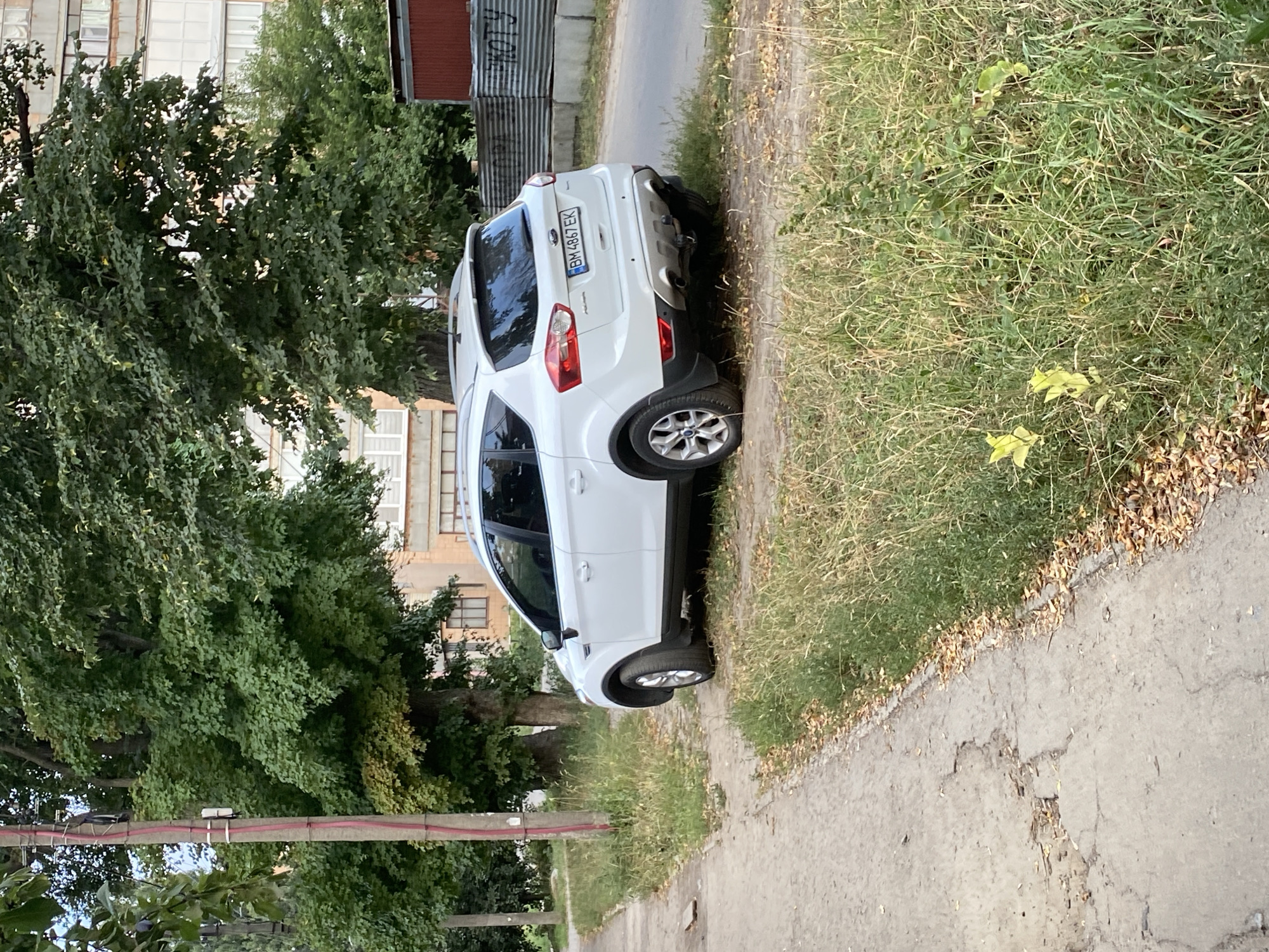 A white Ford SUV is parked on the side of a paved road, partially on a grassy area. The vehicle has a license plate that reads "BH 4867 EK". The scene is an outdoor setting with mature trees and a building in the background. The building appears to be residential or office space with multiple windows. The weather seems to be clear and the time of day is daytime, with shadows visible. The overall impression is a quiet, urban or suburban street. No people are visible in the image.