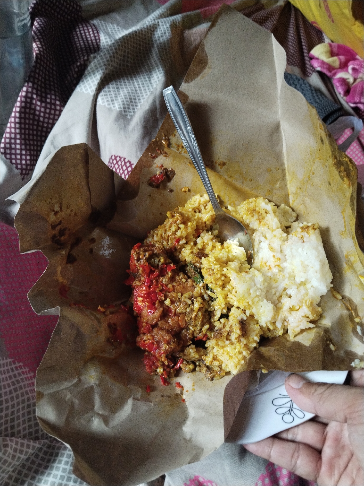 In Cemplang, Indonesia, a close-up, top-down shot captures a meal wrapped in brown paper. The meal consists of rice and a spicy-looking dish with chili peppers and meat. A silver spoon is nestled into the rice, indicating it is ready to be eaten. The food is placed on a surface covered with a patterned fabric, possibly a bed or sofa, with shades of grey, white, and burgundy. The scene is illuminated by ambient light, creating soft shadows. A hand is visible in the lower right corner, holding a small white object with a floral design, partially obscuring the brown paper. The overall presentation suggests a casual, home-style meal.