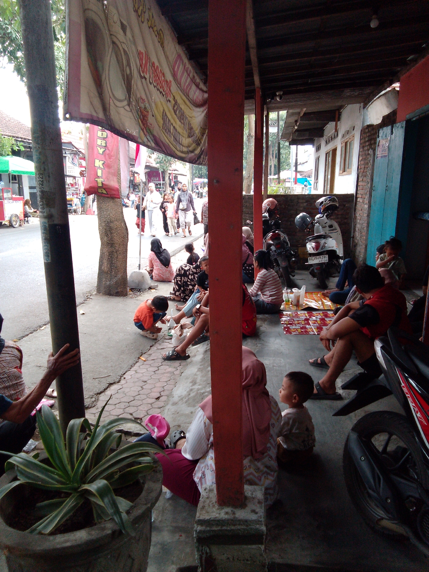 The image depicts a street scene in Trenggalek, Indonesia, likely outside a shop or eatery. A group of people, including adults and children, are gathered on the sidewalk and under a covered area. Some individuals are seated on mats on the ground, suggesting a casual gathering or break. In the background, a street with a few motorbikes and a small cart can be seen. A large banner hangs over the scene, adding to the urban atmosphere. The presence of motorbikes indicates common transportation in the area. The overall impression is a snapshot of daily life in a bustling Indonesian city.