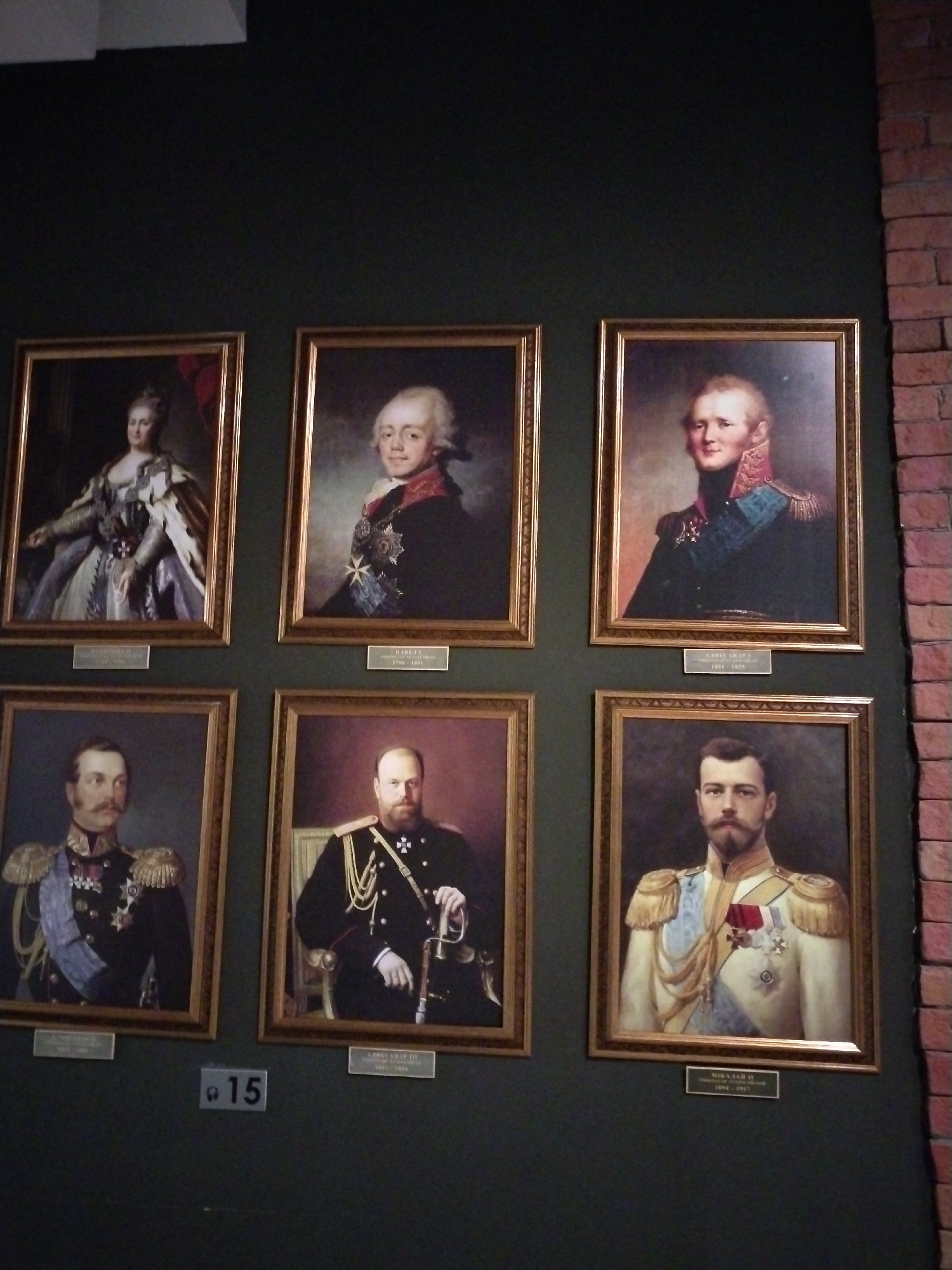 This is a photograph of a display of six historical portraits hanging on a dark green wall. The portraits are arranged in two rows of three. Each portrait is in a golden frame and depicts a single male or female figure in formal attire, likely royalty or military leaders, adorned with medals and military uniforms. Below each portrait is a small plaque with text, presumably identifying the subject and their dates. A brick wall is visible on the right side of the image. There is also a sign with the number "15" in the lower center of the image. The lighting suggests an indoor environment, possibly a museum or gallery. The location is indicated as Камянюцкі сельскі Савет, Belarus.
