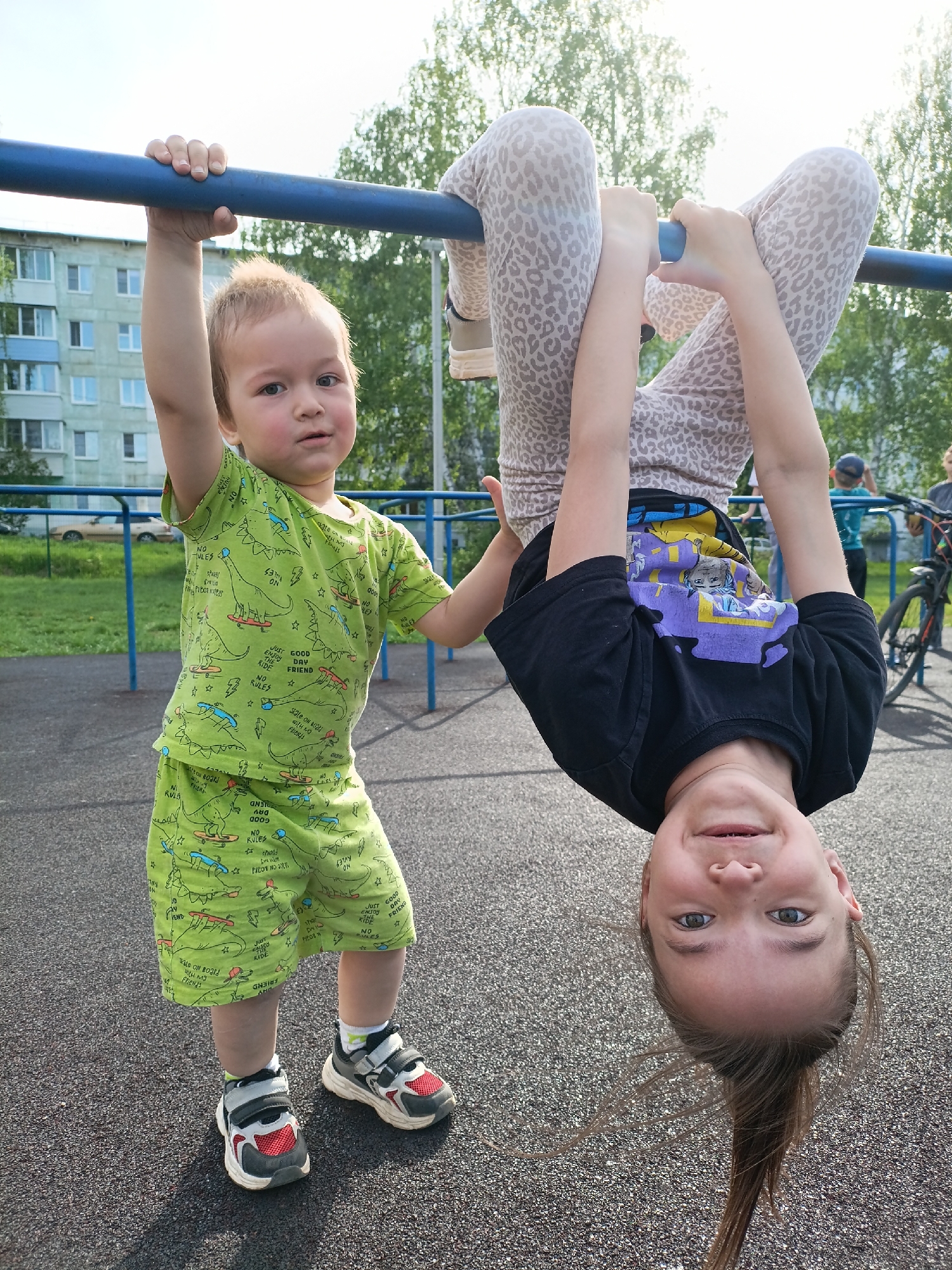 Two children are on playground equipment. One child, a young boy, stands on the ground. He is wearing a green patterned t-shirt and shorts set, and gray and red velcro sneakers. He is holding the blue metal bar of the equipment. Another child hangs upside-down from the bar. She wears animal print leggings and a black t-shirt. Her hair is hanging down as she smiles at the camera. In the background is a multi-story residential building. The image was taken in the city of Chaikovsky, Russia.