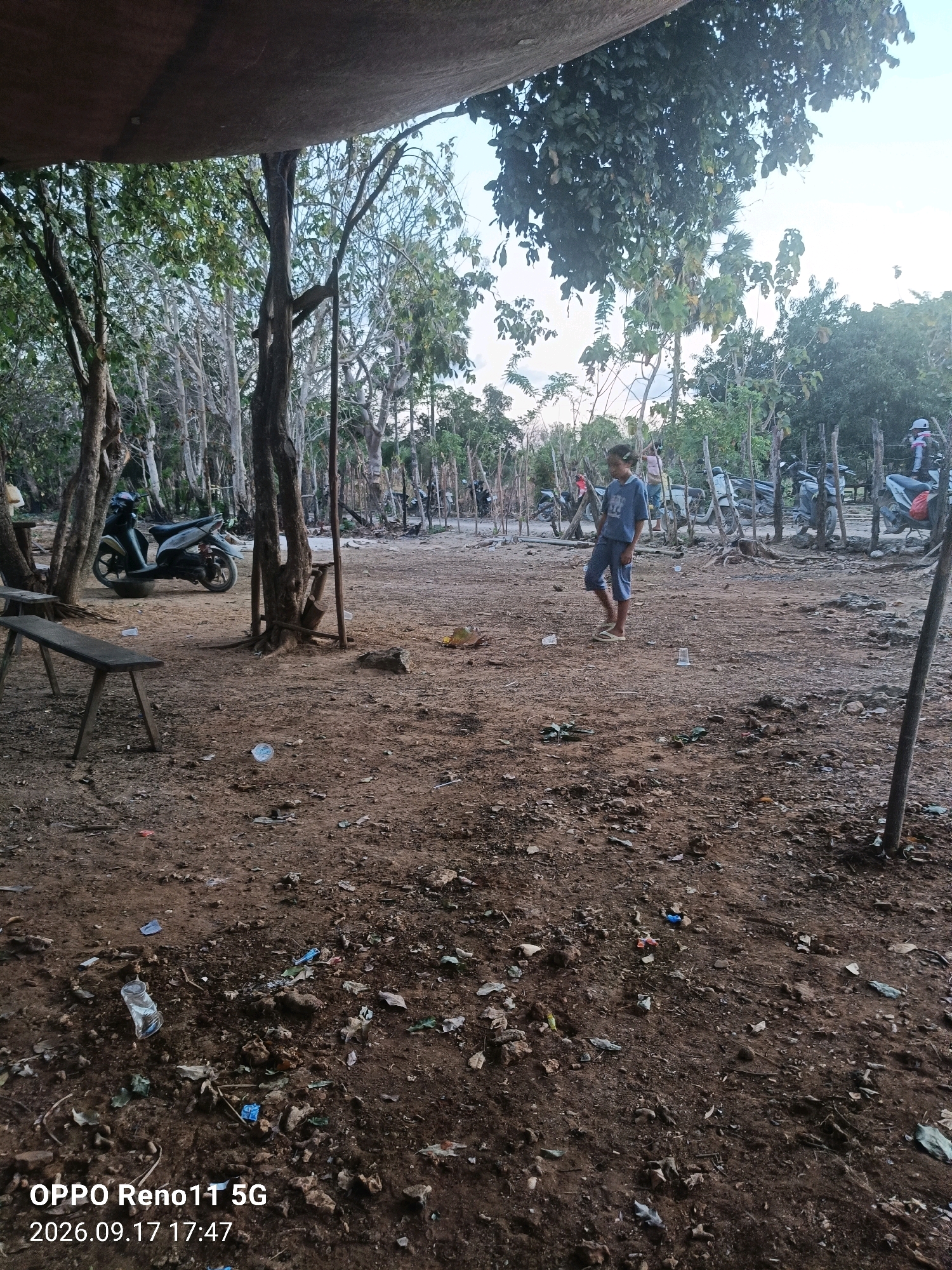 The image depicts an outdoor scene in Otan, Indonesia. In the foreground, a dirt ground is littered with various debris, including plastic bottles and wrappers, leaves, and small stones. A wooden bench is positioned on the left side of the frame. In the middle ground, a young person wearing a blue t-shirt and shorts stands looking to their left. Behind them, several motorcycles are parked. A thin wooden fence runs through the middle ground, separating the open area from a more densely wooded area in the background. The background consists of trees with sparse foliage and a light sky. A dark, tarp-like structure is visible at the top of the frame, casting a shadow over the scene. The overall impression is one of a somewhat unkempt, rural or semi-rural environment.