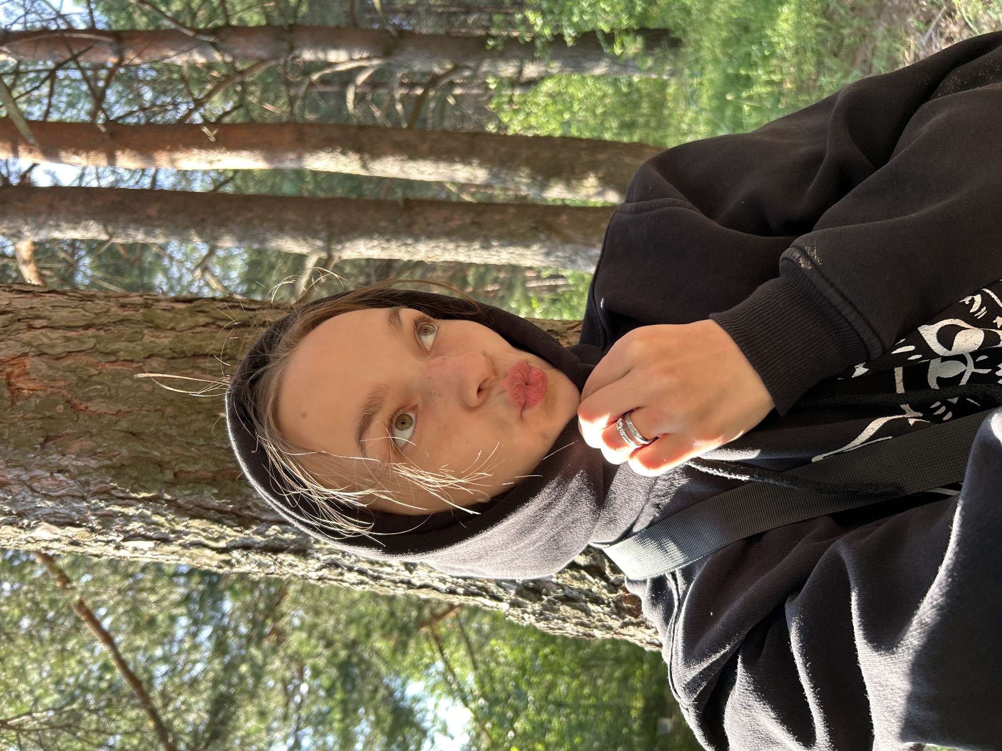 A young person with light brown hair is leaning against a tree, looking towards the camera with a playful expression and puckered lips. They are wearing a black hoodie with the hood pulled up, partially obscuring their hair. A silver ring is visible on their right index finger. The background consists of a forest with tall trees and dappled sunlight, suggesting an outdoor setting during the day. The image appears to be taken in Chaikovsky, Russia. The overall mood is casual and lighthearted.