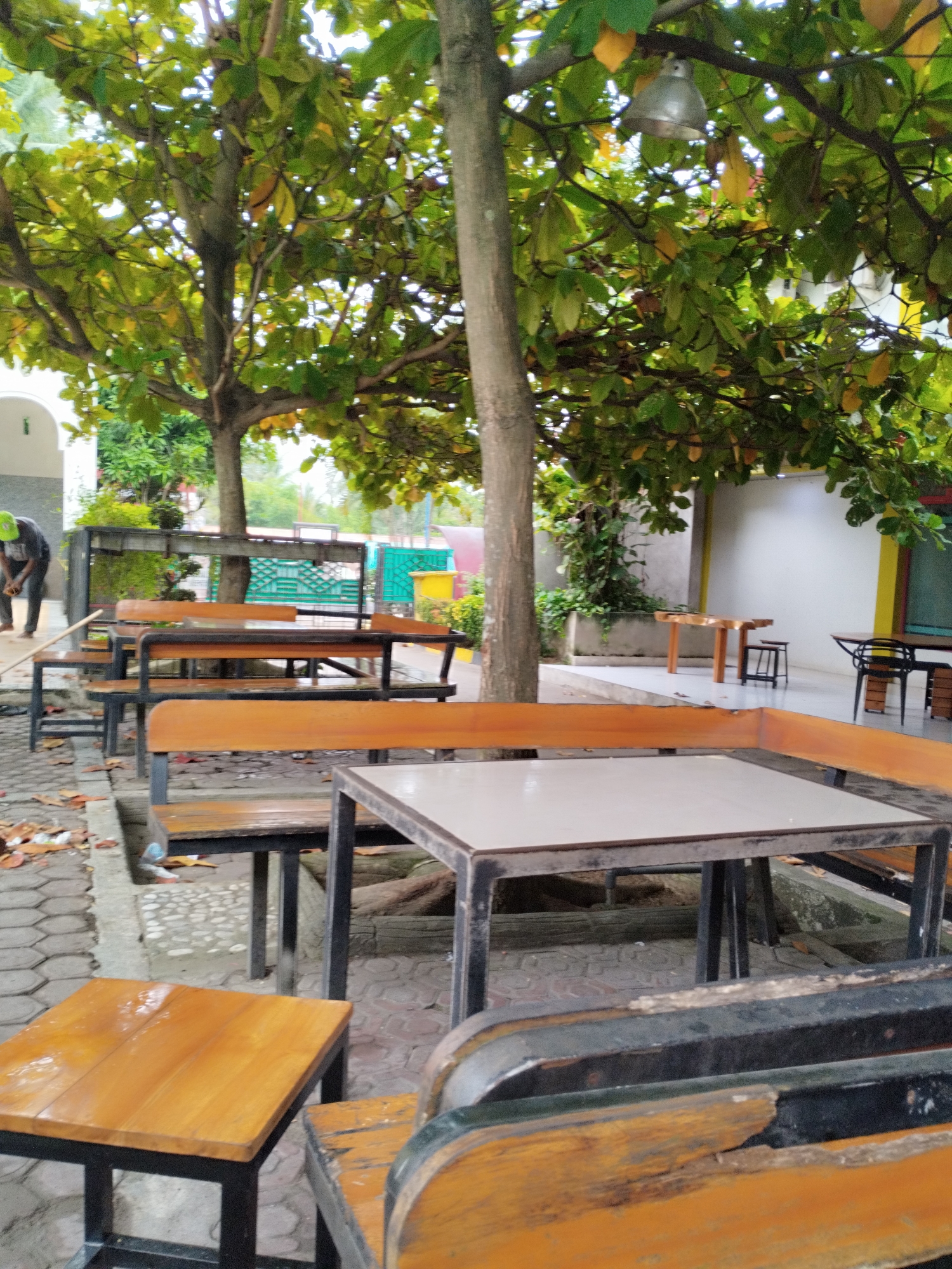 The image captures an outdoor seating area with wooden tables and benches beneath a leafy tree. The ground is paved with hexagonal stones, and fallen leaves are scattered around. In the background, there is a white wall with some green fencing and a glimpse of other outdoor furniture. A person wearing a green cap and dark clothing is visible in the far left of the image, appearing to be engaged in some activity on the ground. The scene suggests a casual, possibly cafe or restaurant setting. The presence of trees and the outdoor arrangement indicates this location is in an environment with a tropical or subtropical climate. This scene is situated in Payameuneng, Indonesia.