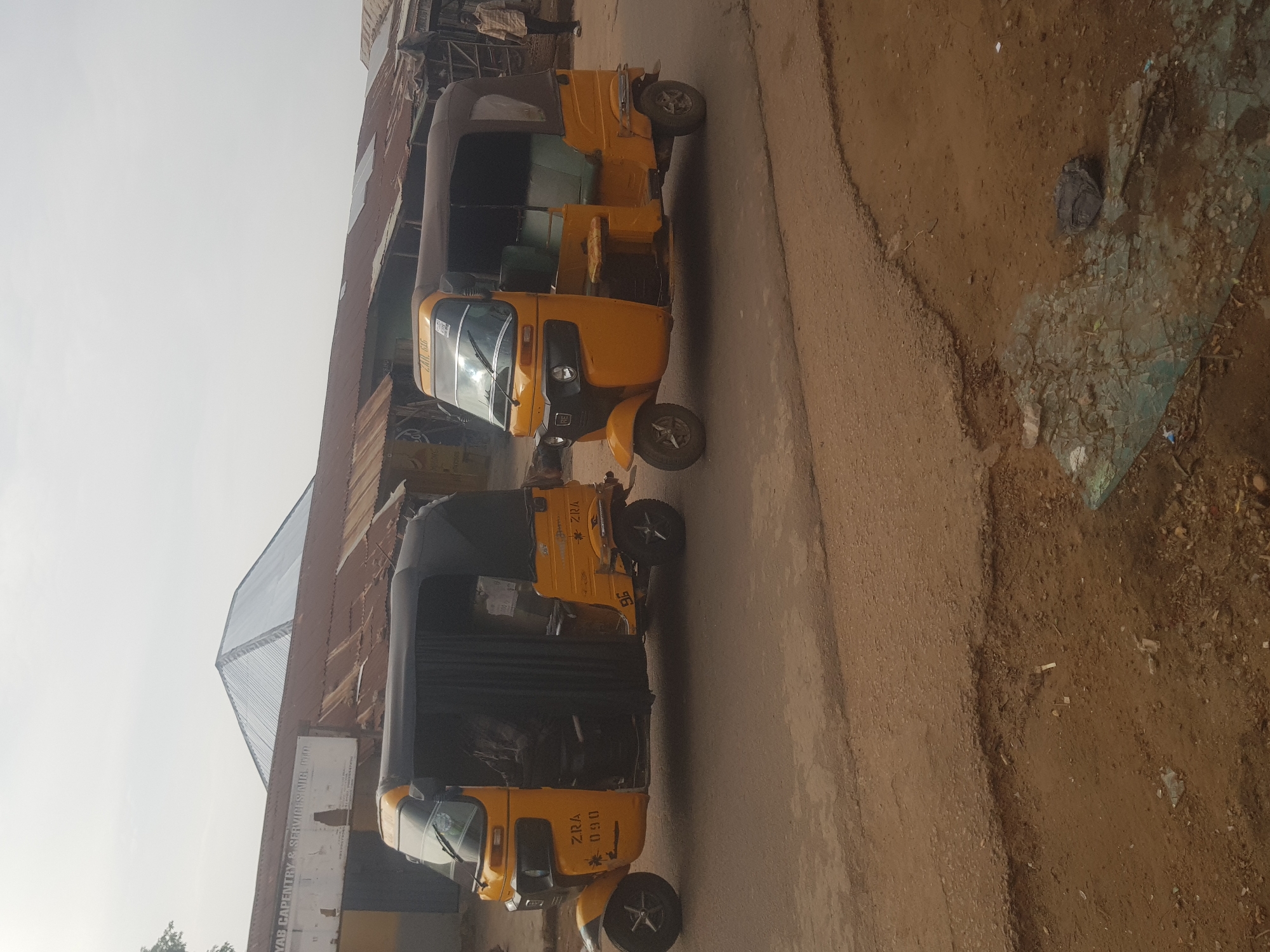 The image depicts two yellow and black "Keke Napep" auto-rickshaws parked on a paved road. The rickshaws are adorned with "ZRA" lettering. The background features a building with a metal roof and a sign for "ZYAB Carpentry & Services Nigl Ltd." The surrounding environment appears to be a dusty urban roadside in Tukur Tukur, Nigeria. There are no visible people in the immediate scene.