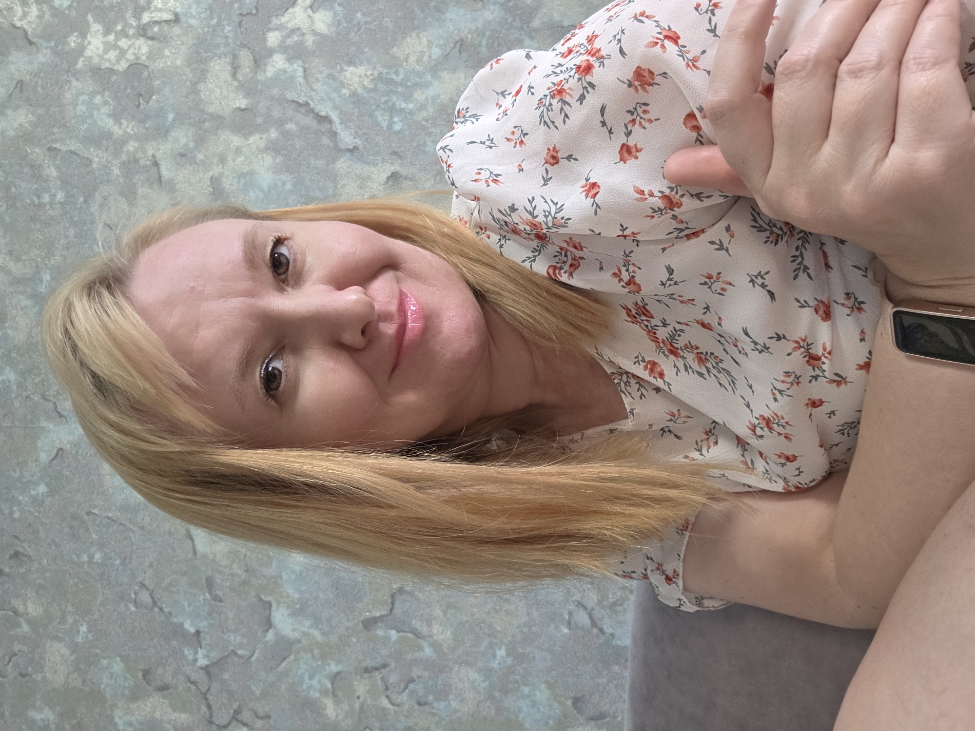 A blonde woman with fair skin and light makeup is pictured from the chest up, seated on a gray sofa. She is wearing a white blouse with a floral pattern of red and green flowers. Her arms are crossed, with her left arm resting on top of her right. Her hair is shoulder-length and blonde. She is looking directly at the camera with a slight smile. The background is a textured gray and blue wall. The scene appears to be indoors, in a residential setting.