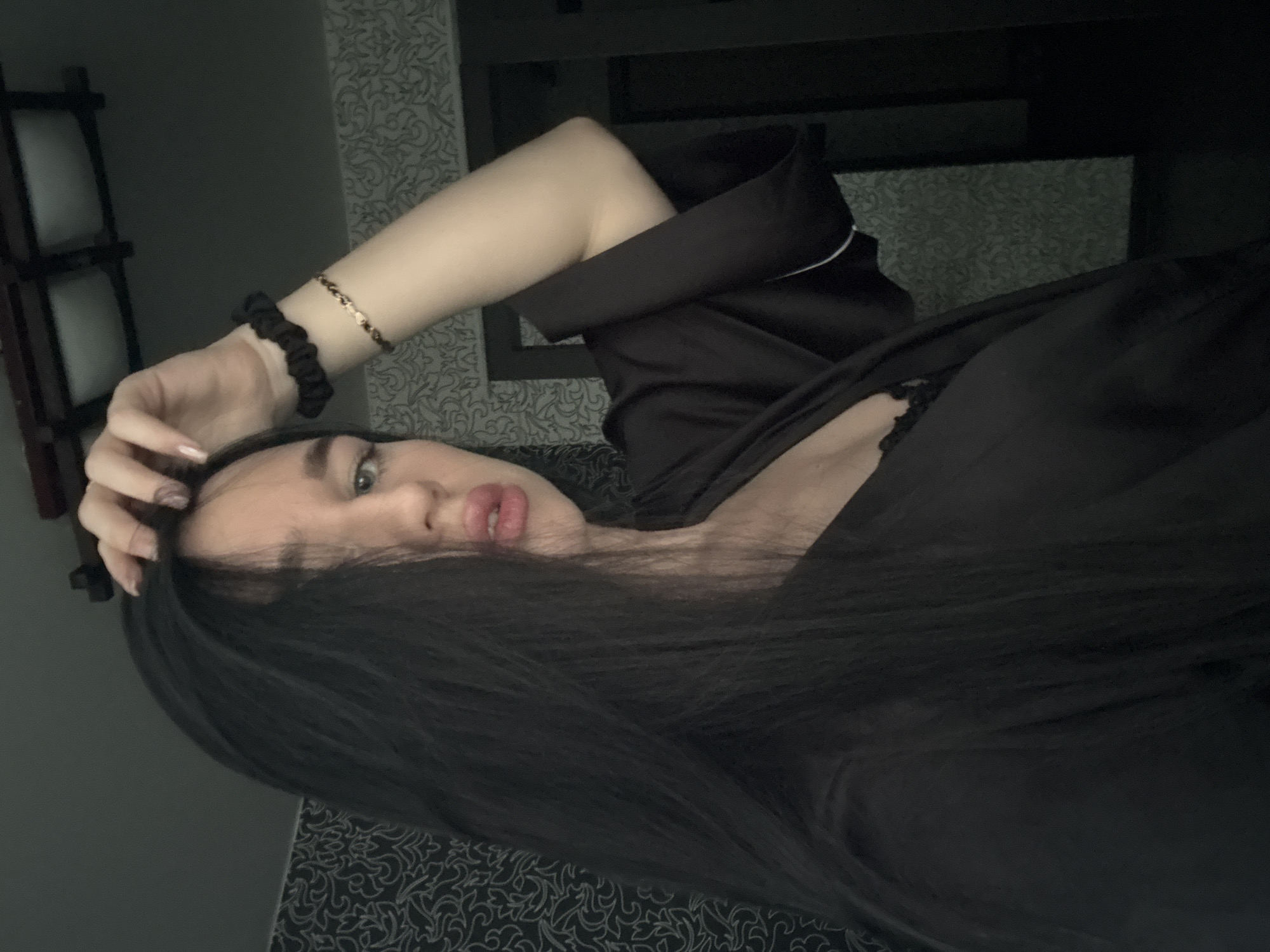 A young woman with long, dark hair and light skin is pictured in a close-up shot. She is wearing a black satin robe, revealing a black lace bra underneath. Her left hand is resting on her head, and she is wearing a black scrunchie and a thin gold bracelet on her left wrist. The background appears to be an indoor setting with patterned wallpaper and a portion of a doorway or closet visible. The lighting is dim. There is no discernible information in the image to suggest the location is Novosibirsk, Russia.