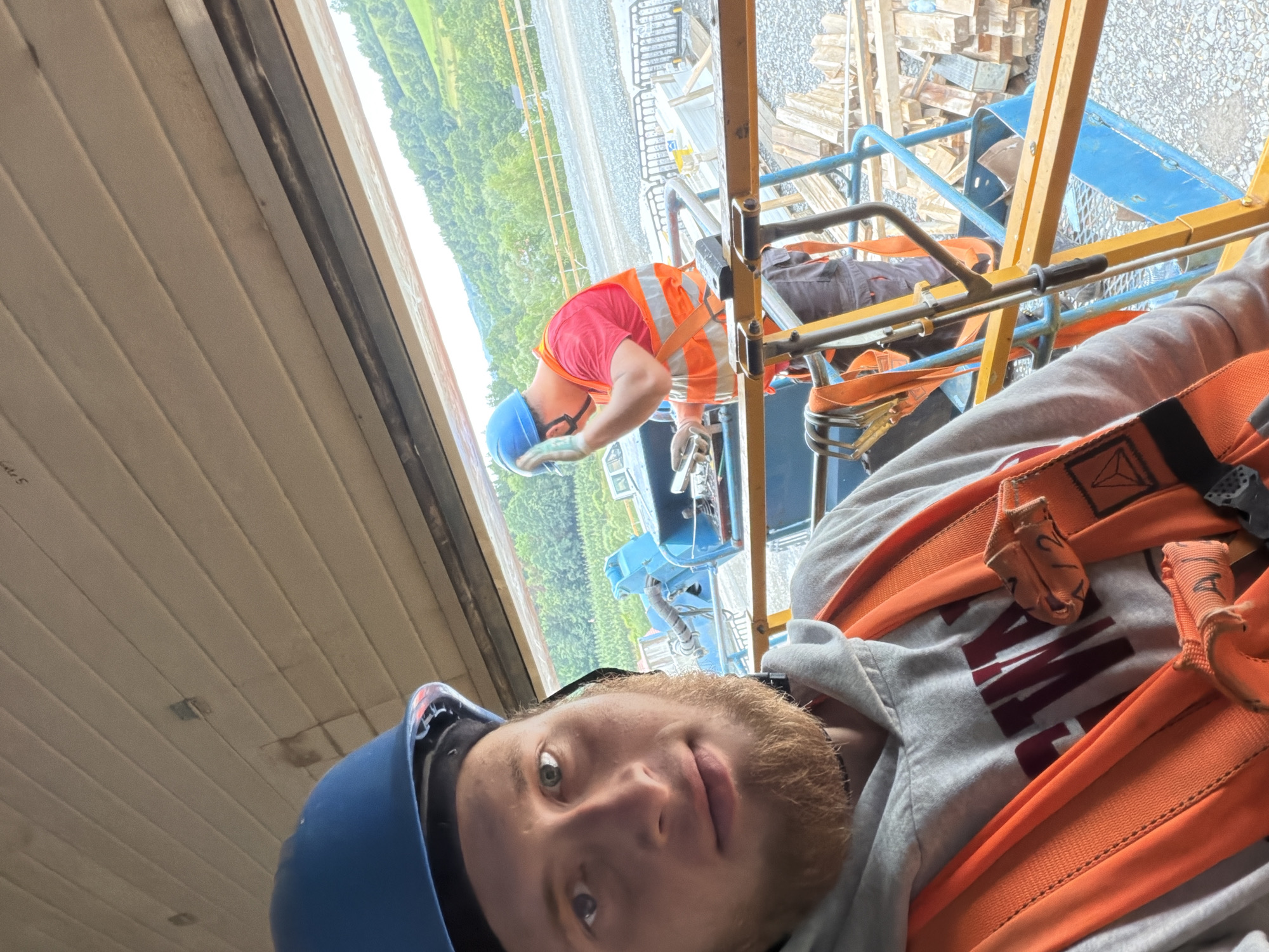 This media file shows two construction workers on a scaffolding platform. The main subject is a young man in the foreground, wearing a blue hard hat and a grey hoodie with an orange safety harness. He is looking directly at the camera with a slight smile and has light-colored hair and beard. To his right, partially obscured by his head, is the orange strapping of his harness.
Behind him and to the left, another worker is visible. This worker is wearing a red shirt, an orange safety vest, a blue hard hat, and gloves, and is bent over, appearing to be engaged in work. Orange safety harness straps are also visible on this worker. The scaffolding structure, made of yellow and blue metal poles, is a prominent object in the scene.
The background reveals an outdoor setting with a lush green hillside and trees, suggesting a rural or semi-rural location. There are also piles of lumber and construction debris in the background, indicating an active construction site. The lighting suggests it is daytime. The overall mood appears to be casual, with the foreground subject taking a selfie during what seems to be a break or a moment of downtime on the job. No visible text is legible in the image. The location context is Janowice, Poland.