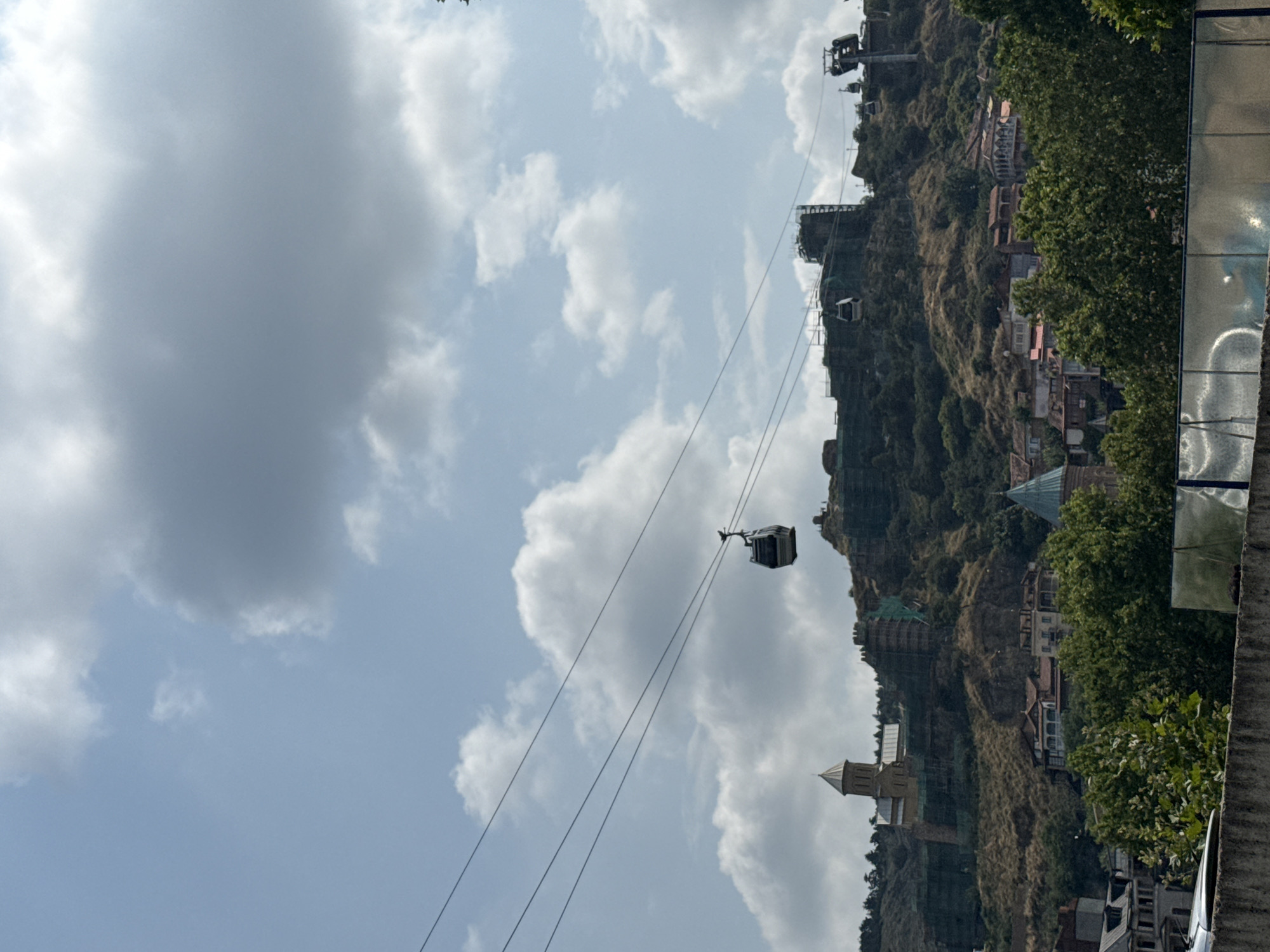 In Tbilisi, Georgia, a scenic view of the city unfolds beneath a sky dotted with clouds. Cable cars traverse the sky on thin wires, with one cabin prominently visible in the middle ground. The cable cars appear to be moving across the frame. The background is dominated by a lush, green hillside dotted with trees and buildings. Among the buildings, a distinct church with a conical roof and a tower stands out. Further up the hill, remnants of a historic fortress or structure, possibly undergoing renovation with green netting, are visible. The lower part of the image shows a concrete barrier and some foliage, suggesting the viewpoint is from a slightly elevated position.