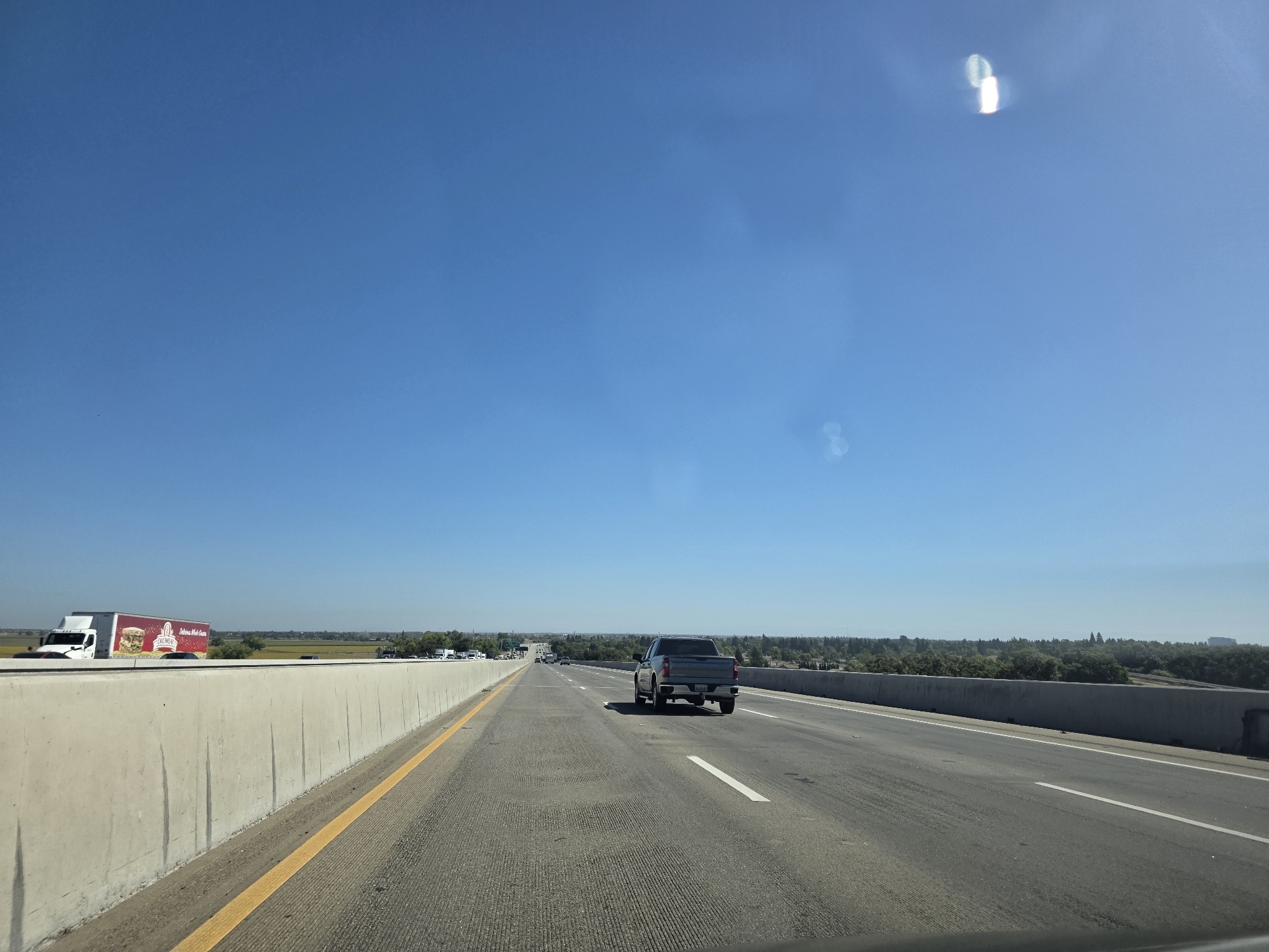 The image captures a view from a vehicle traveling on a highway under a clear blue sky. The road has multiple lanes, with a concrete barrier on the left and a low retaining wall on the right. A white semi-trailer truck with a red and white logo is visible on the left side of the road, and a gray pickup truck is in the right lane ahead. In the distance, trees and buildings can be seen, suggesting a developed area. The time of day appears to be late morning or early afternoon, indicated by the bright sunlight and long shadows. The weather is clear and sunny. The setting is a highway, likely in a rural or suburban area given the presence of trees and open fields beyond the roadside. There is no discernible emotion conveyed by the image, which presents a straightforward, objective view of the environment. The text on the side of the semi-trailer truck is visible, showing a logo with the word "OROWEAT" and an image of bread.