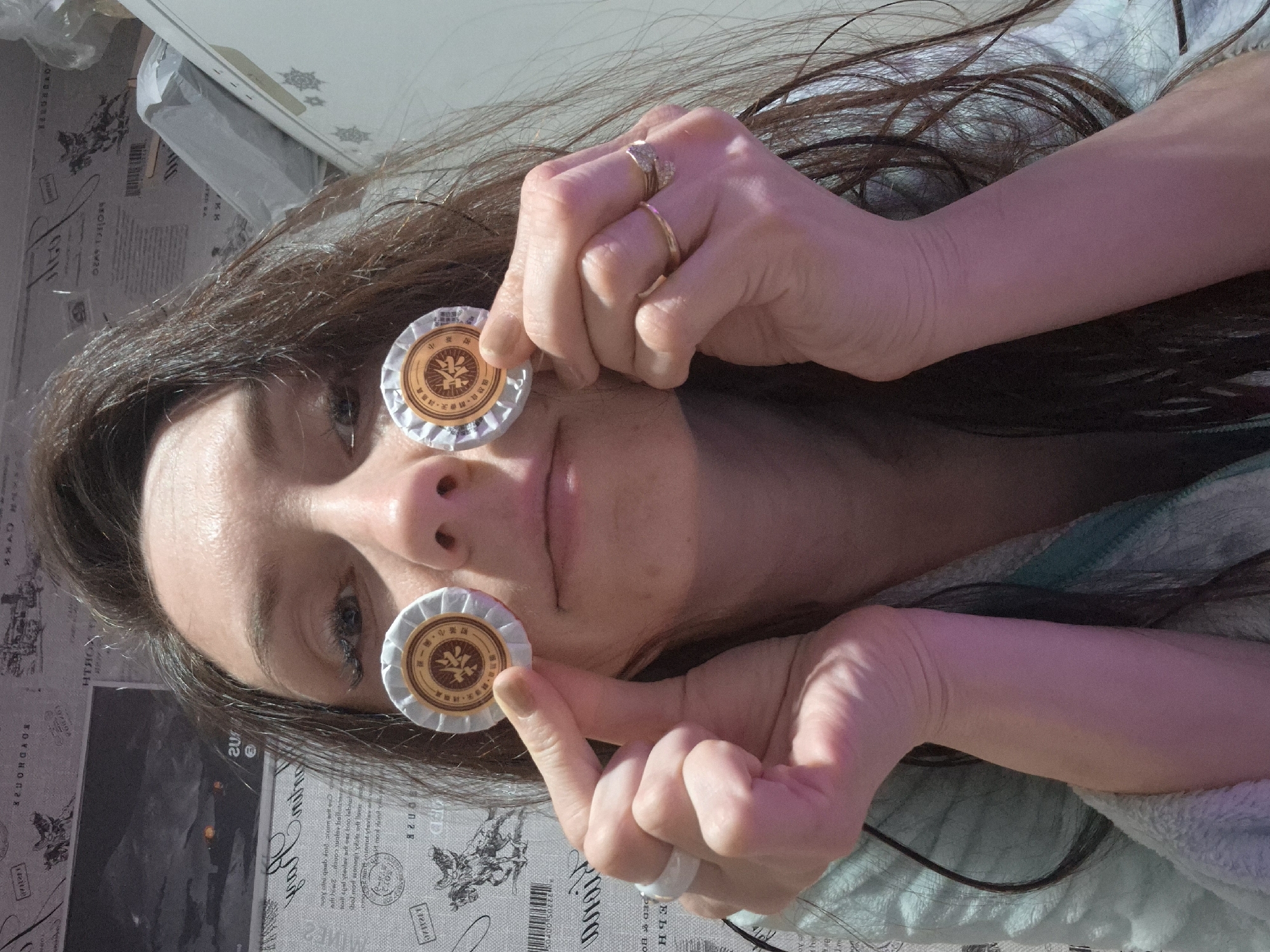 A woman is lying down, holding two small, round tea sachets up to her eyes like makeshift glasses. She has long, dark brown hair, fair skin, and is wearing rings on her fingers. The tea sachets have a beige-colored center with a dark brown logo, surrounded by white crimped edges. The background appears to be a patterned surface, possibly a tablecloth or a covering with black and white drawings and text. The overall lighting is bright and sunny.