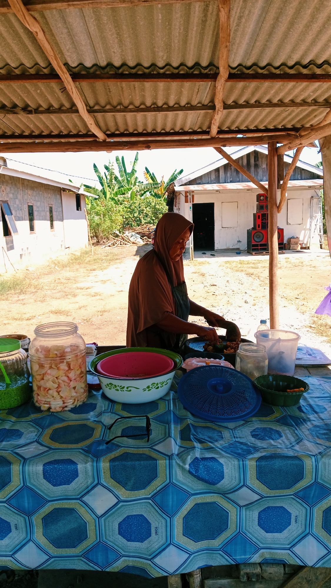 An Indonesian woman wearing a brown hijab and an apron is standing behind a table under a simple wooden and corrugated metal roof. She is preparing food with a mortar and pestle on a black pan. The table is covered with a blue and white patterned tablecloth and holds various containers of food, including a large glass jar filled with sliced fruit, two bowls, a blue lid, and a green bowl with sliced carrots. In the background, there are simple buildings, including one made of cinder blocks and another with a wooden exterior. Banana trees and other vegetation are visible. Two large speakers are stacked against the side of one of the buildings. The scene appears to be an outdoor food stall in a rural or semi-rural area of Karang Bunga, Indonesia, on a bright, sunny day.