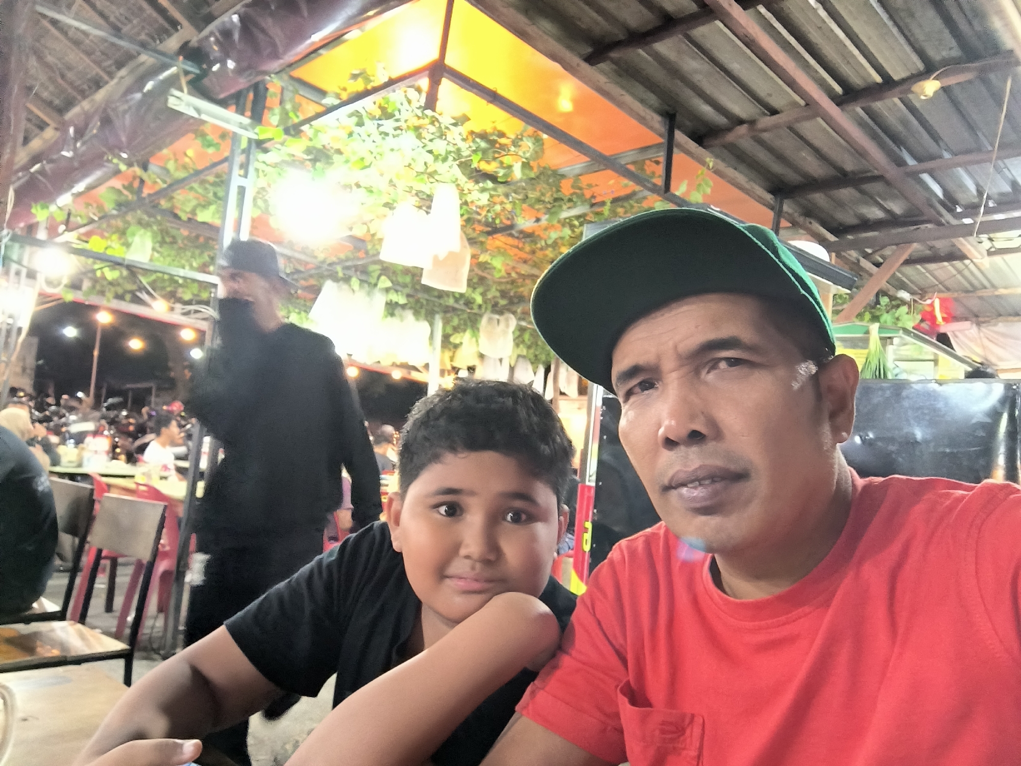 The image is a close-up selfie taken at night, featuring an adult man and a young boy, likely in an informal outdoor eatery or night market in Banda Aceh, Indonesia. The man, positioned on the right, wears a vibrant red t-shirt and a green baseball cap, looking towards the camera with a neutral and observant expression. To his left, a young boy in a black t-shirt rests his chin thoughtfully on his hand, his eyes wide and looking directly at the camera with a slight, gentle smile.
Behind them, the scene is lively and blurred, indicating a crowded environment illuminated by numerous artificial lights, confirming the nighttime setting. An overhead structure provides cover, featuring a wooden plank roof interspersed with translucent orange panels that emit a warm glow. Green foliage, possibly artificial vines or plants, is draped across this structure, alongside several hanging white, bag-like objects that serve as decorations. In the mid-ground, glimpses of tables, chairs, and other indistinct figures can be seen, suggesting a casual and bustling atmosphere typical of a street food area. No discernible text is visible in the photograph.