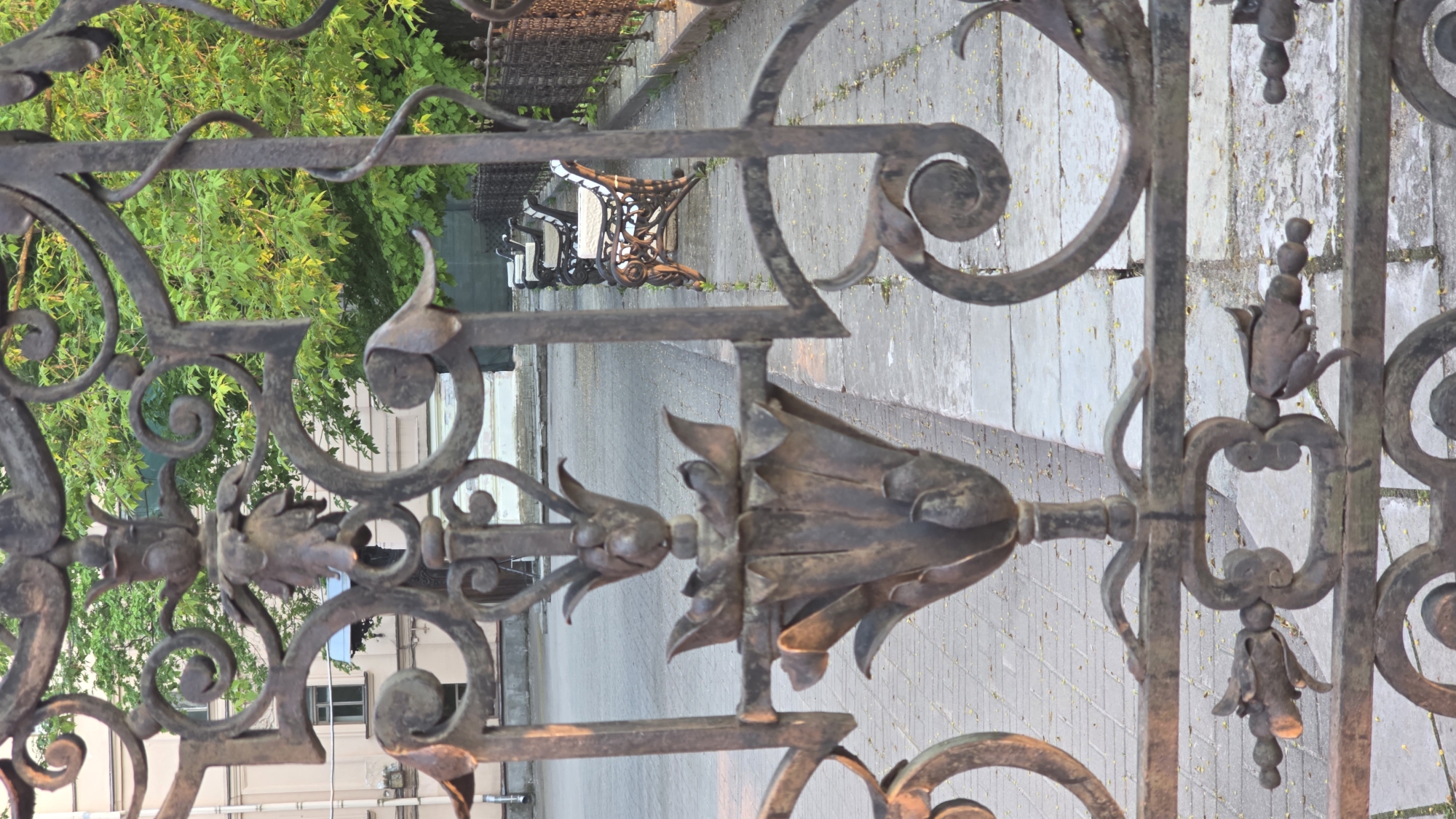 A wrought iron gate in Saint Petersburg, Russia, with floral motifs is centered. Behind the gate, a paved walkway stretches into the distance. A wrought iron park bench is visible along the walkway. Lush green trees grow on the left side of the frame. Buildings are visible in the lower left.