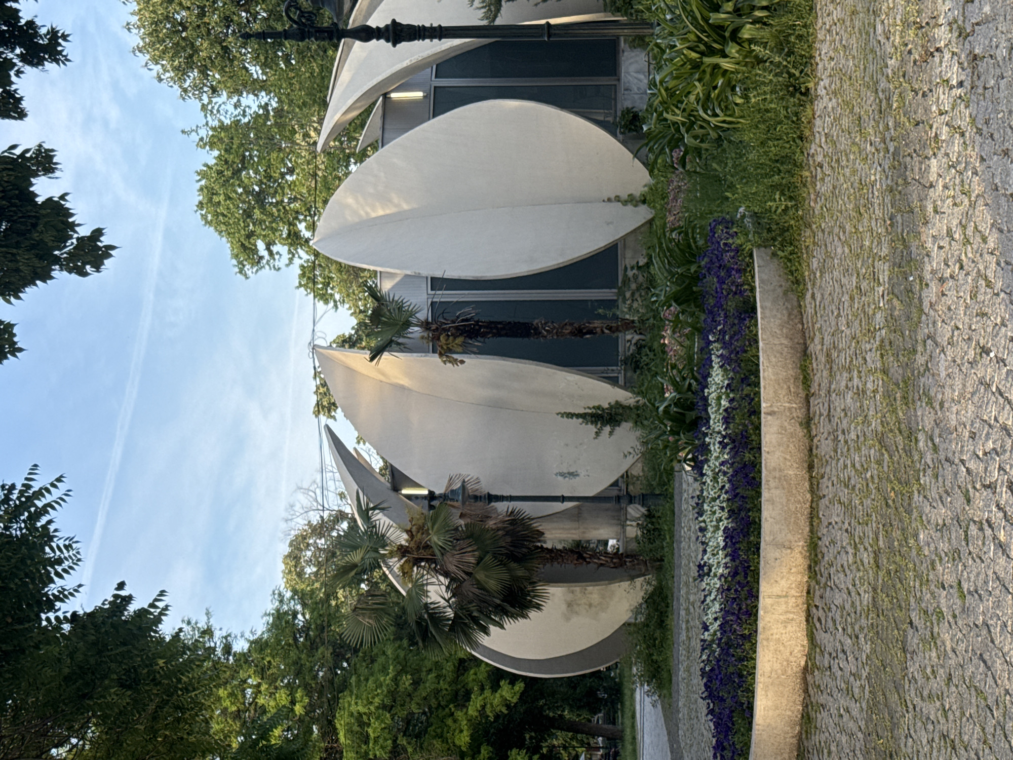 The image shows the "Leaves of the Trees" monument in Plovdiv, Bulgaria. The monument consists of abstract, stylized white structures resembling leaves. These are arranged in a cascading fashion and are complemented by surrounding landscaping, including flowers and trees. A palm tree grows in front of the leaves. A paved stone walkway runs along the right side of the image. The sky is visible through the surrounding trees.