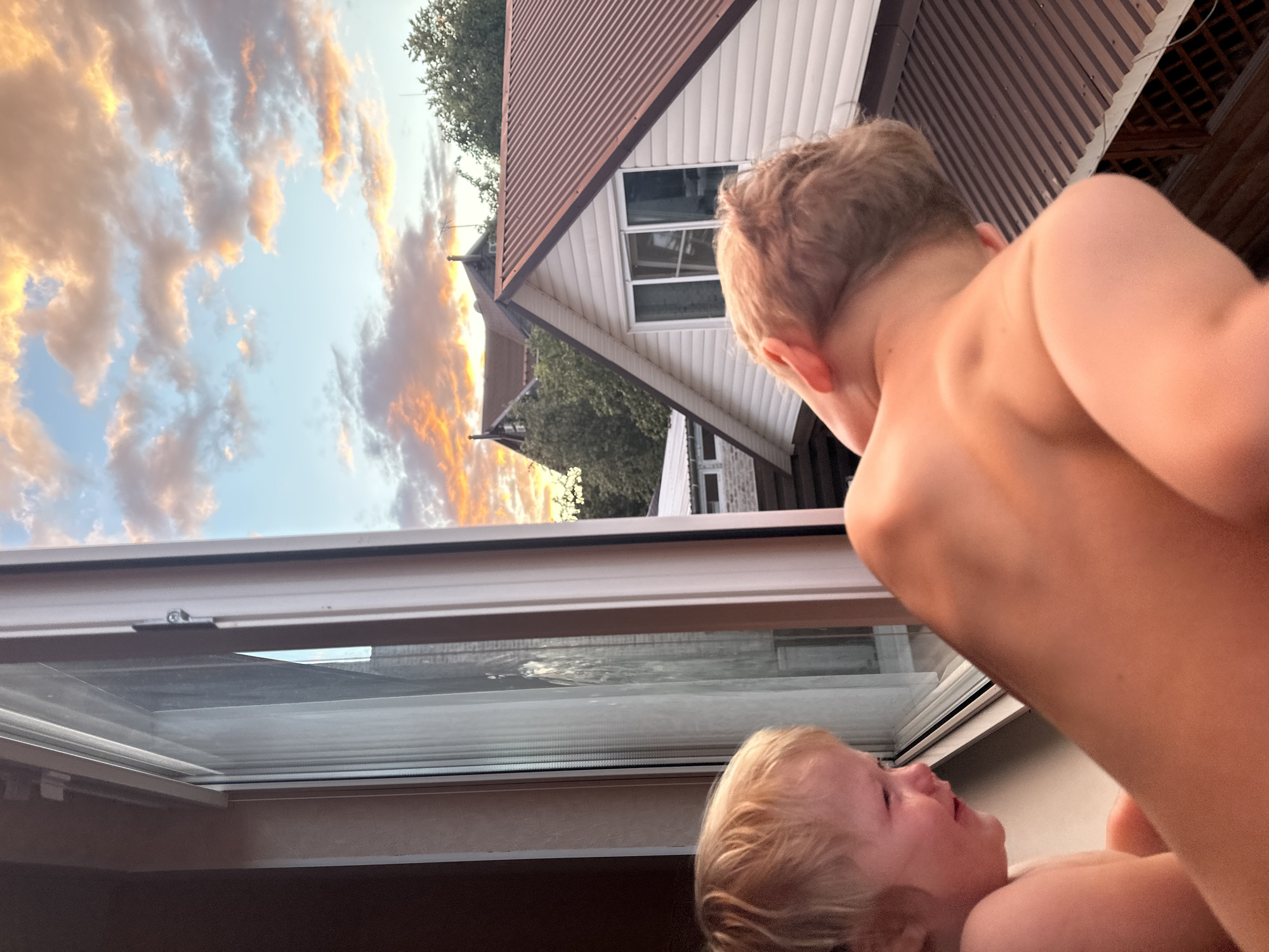 The image shows two young children looking out a window at a sunset. The sky is filled with dramatic clouds, tinged with orange and yellow. In the background, a house with a brown metal roof and white siding is visible. The setting appears to be a residential area in Nizhny Novgorod, Russia. The lighting suggests it is late afternoon or early evening. The children's faces are not clearly visible, but their posture suggests curiosity and wonder.