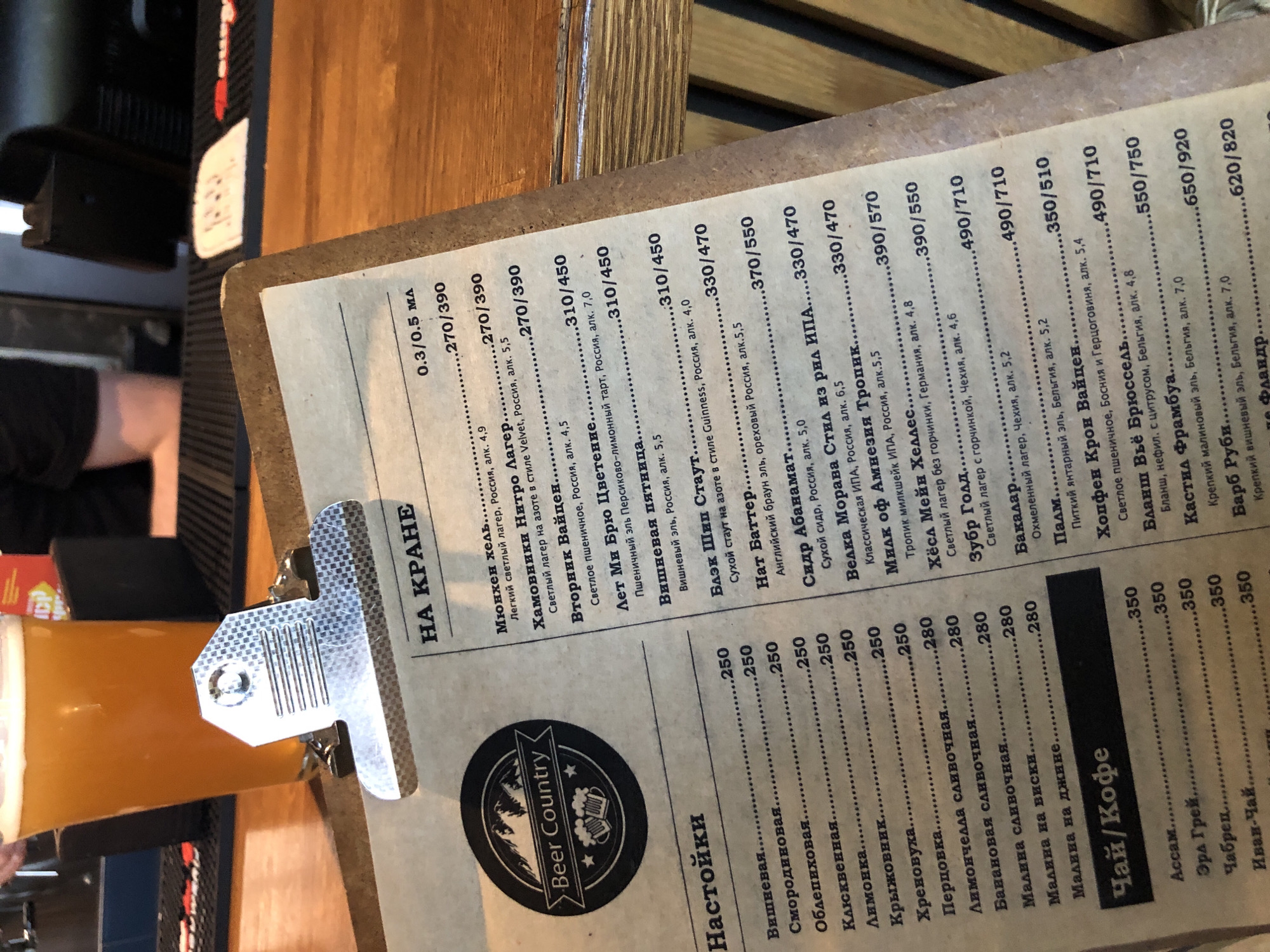 The image shows a close-up view of a menu on a clipboard, resting on a wooden table in front of a partially visible glass of amber liquid, likely beer, with a foamy head. The menu is printed on textured, light brown paper and features a logo on the left side that reads "Beer Country" with an illustration of mountains and beer mugs. The menu itself is divided into sections. The top section, titled "НА КРАНЕ" (On Tap), lists various beers with their descriptions, origins, alcohol content, and prices in Russian. Below this is a section titled "Настойки" (Tinctures/Infusions), listing different flavored infusions with prices. Further down, there is a section labeled "Чай/Кофе" (Tea/Coffee) with a list of teas and their prices. The overall lighting suggests an indoor setting, possibly a bar or restaurant. The background shows a blurry bar counter and what appears to be a person's legs. The focus is on the menu, with the clipboard and the beverage serving as foreground elements.