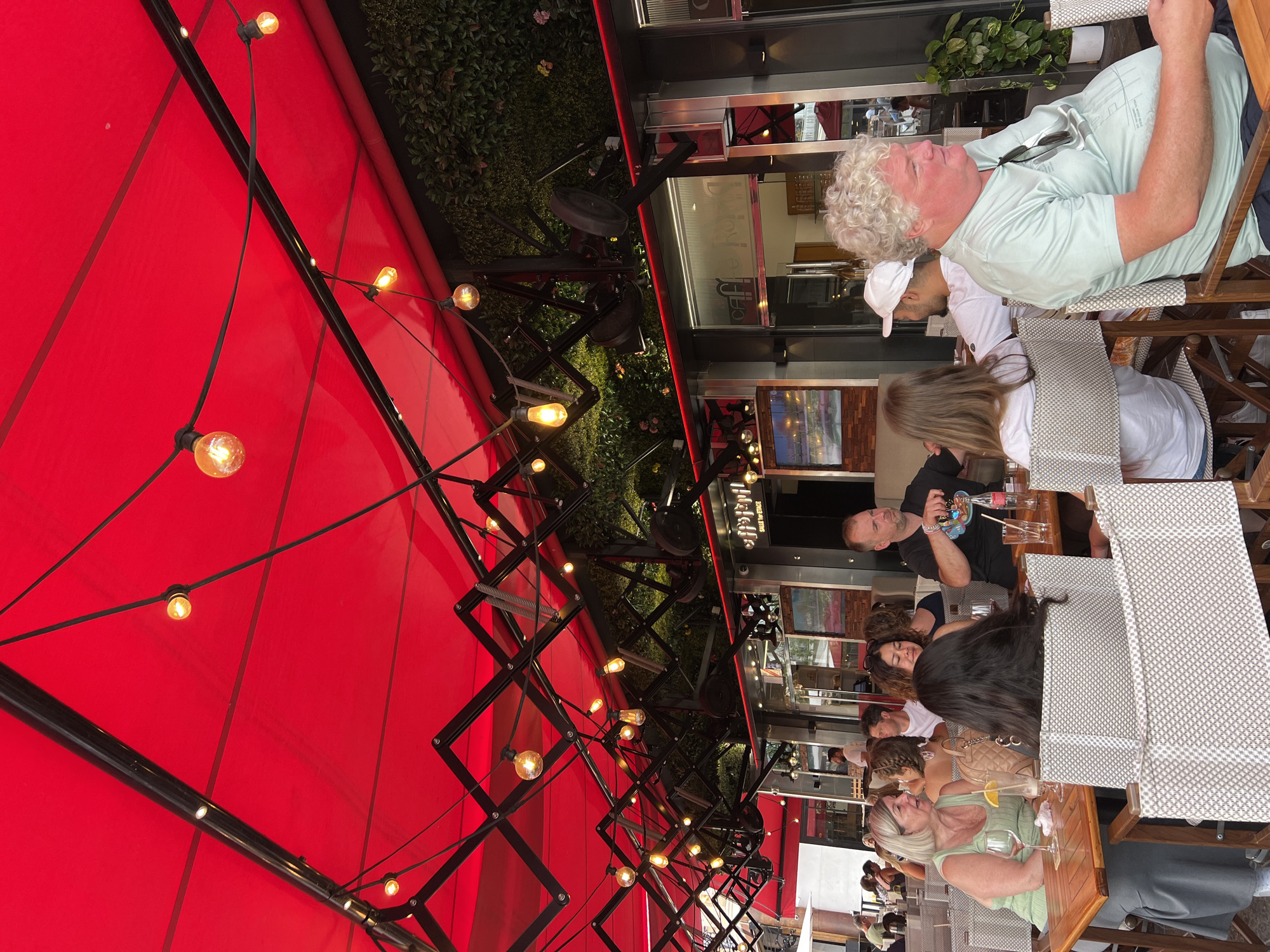 This image depicts an outdoor dining area of a restaurant, likely located in Cannes, France, under a bright red awning adorned with string lights. Several groups of people are seated at tables, engaged in conversation and dining. In the foreground, a woman with blonde hair wearing a green top is seated at a table with a drink. To her right, another woman with dark hair is seated facing away from the camera. A man in a black t-shirt is seated at a table further back, holding a bottle. In the mid-ground, a group of diners are visible, including a man in a white t-shirt and a white cap. The background shows the restaurant facade with large windows and a green wall covered in plants. The overall atmosphere suggests a lively and pleasant dining experience in a city setting.