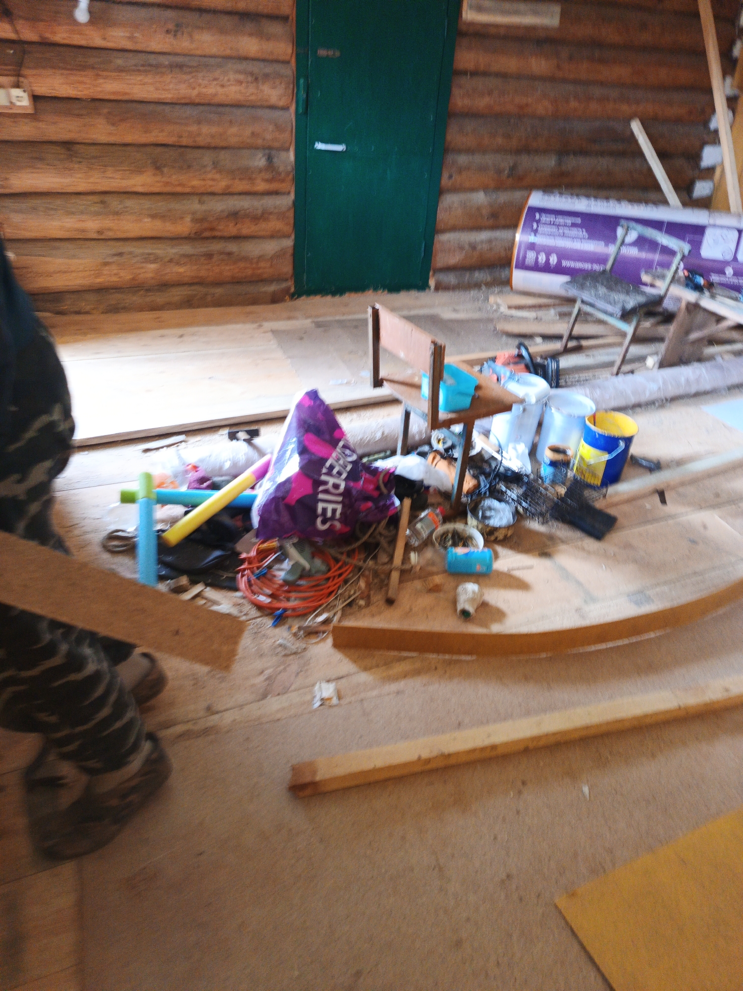 The image depicts an indoor scene, likely a workshop or storage area, within a wooden log structure. The walls are constructed from horizontal logs, and a dark green door is visible on the left side. The floor is wooden, covered in sawdust and debris.
In the foreground, a person's legs are visible, wearing camouflage-patterned pants and dark boots. They are standing near a piece of light-colored lumber.
The midground is cluttered with various objects. To the left, there are colorful items like blue and yellow foam pool noodles, and a purple bag with the word "DEERIES" visible. Various cables, including an orange coiled one, and other miscellaneous items are scattered around. Further back, there's a chair made of wood and metal, and several buckets, one yellow and blue, and others white. A large purple-wrapped cylindrical object, possibly insulation or a roll of material, leans against the wall. Some construction materials, like planks of wood, are stacked or leaning.
The background shows more of the log wall and a section of what appears to be a white ceiling or higher wall. The overall impression is one of a work in progress or a disorganized storage space. The image quality is somewhat blurry, making precise identification of all objects difficult. The location is specified as Islambaevo, Russia.