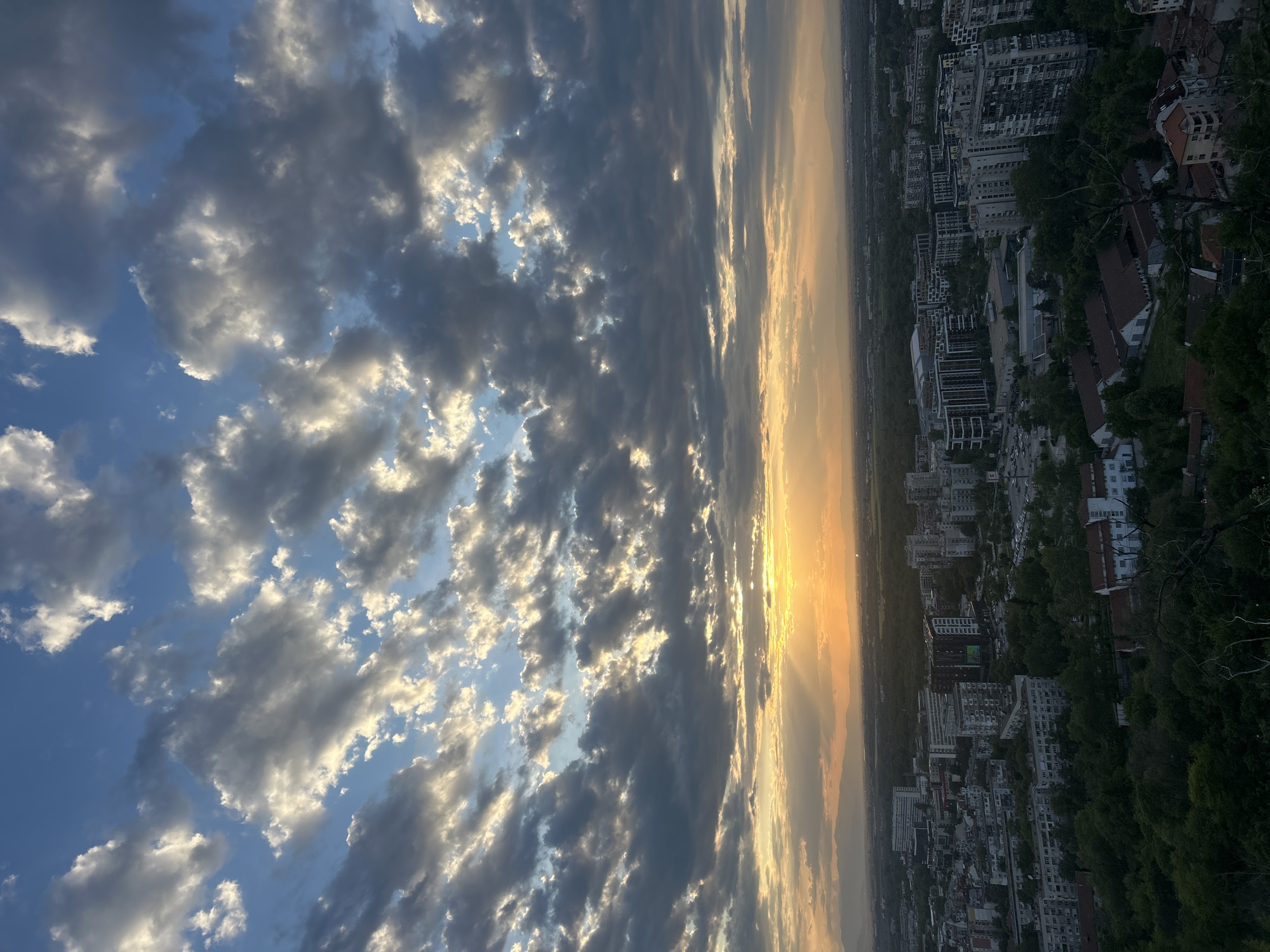 The image captures a sunset view over Plovdiv, Bulgaria, from what appears to be a high vantage point. In the distance, the city's buildings stretch along the landscape under a sky filled with textured clouds. The sunset casts a warm golden glow on the horizon, where the sky is less cloudy. The angle of the photo suggests it was taken looking towards the horizon from a height, potentially a hill or building overlooking the city. Trees and other vegetation are visible in the foreground, framing the urban panorama.