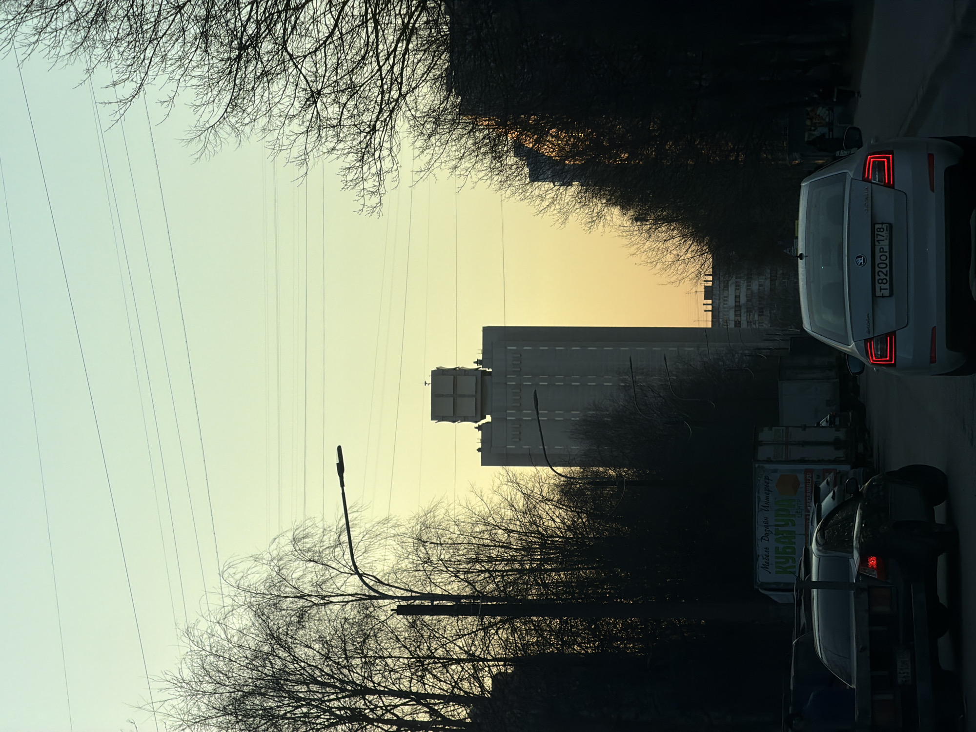 The image depicts a street scene in Saint Petersburg, Russia, at what appears to be dusk. The sky is a pale yellow-orange, indicating sunset. A tall, light-grey building is visible in the middle distance, partially obscured by leafless trees. Power lines are strung across the sky. On the right side of the frame are two cars, one light-colored and the other dark. The license plate of the light-colored car reads "T 820 OP 178", which is a Russian vehicle registration. A furniture advertisement is also visible on the right. Leafless trees frame both sides of the image. A streetlight is visible on the left side of the frame. The image is oriented sideways.