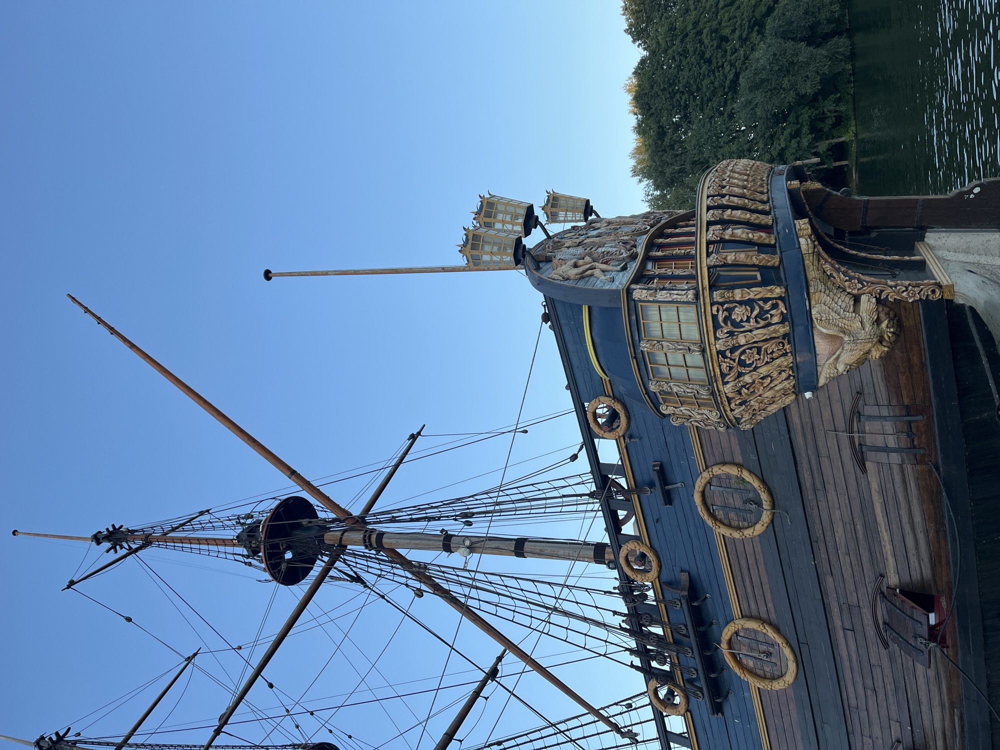 This is a full, eye-level shot of a historic sailing ship docked on a body of water. The ship, painted dark blue and brown, features elaborate golden carvings on its stern and along its railing. Tall masts and rigging dominate the left side of the frame, with a clear blue sky as the background. Trees are visible on the right bank of the water, suggesting a park or natural setting. The ship's name, "Gostiny Dvor," is visible on the side. The ship is located in Novaya Usman, Russia. There are no people visible in the image.