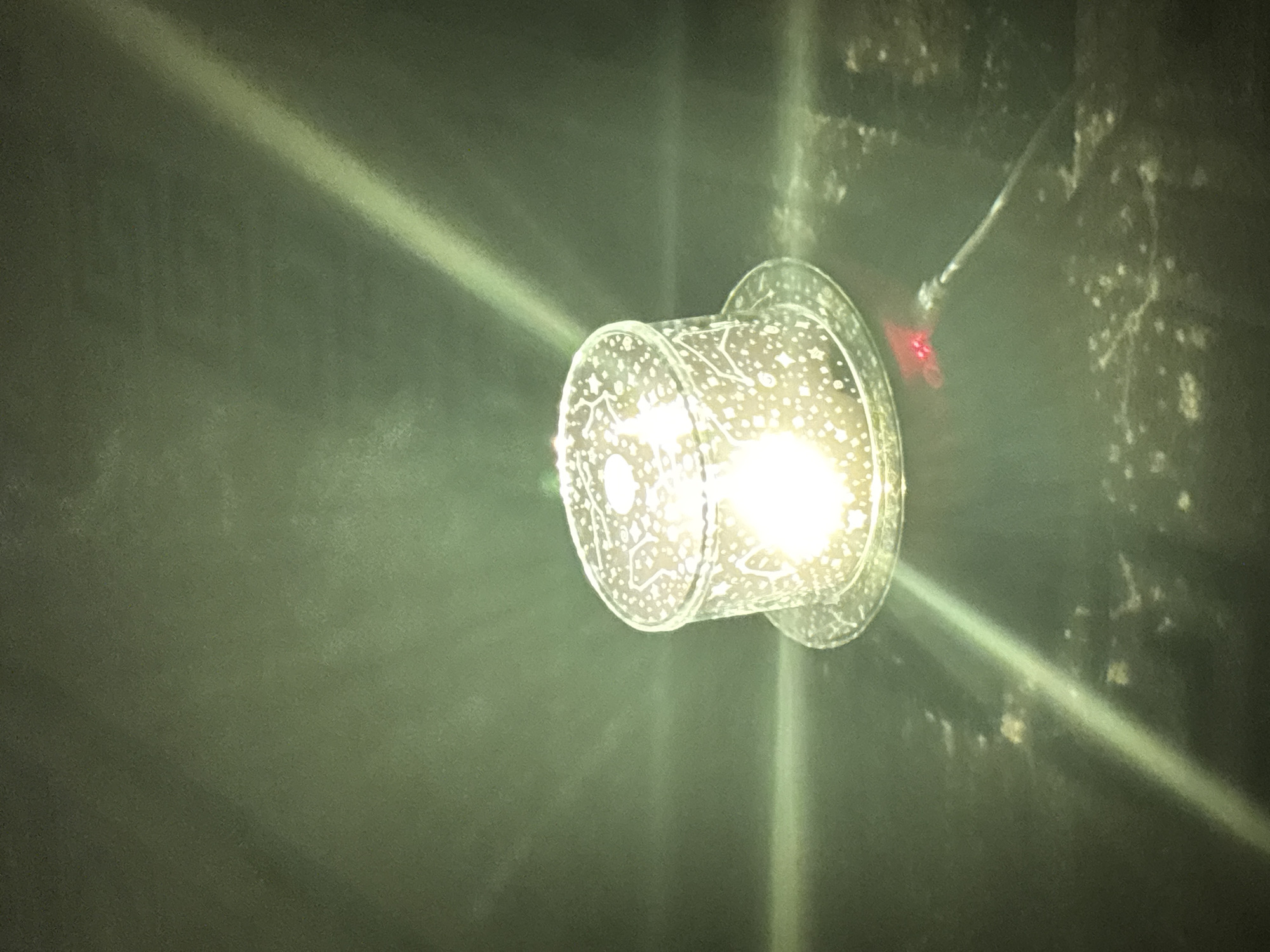 A clear cylindrical glass object with a clear flat base, possibly a candle holder or diffuser, is illuminated from within by a bright light source. The glass is decorated with a pattern of small white dots and stars. A red LED light is visible on the base of the object, and a dark cable extends from it. The object rests on a dark, textured surface, with light beams reflecting across it. The background is a dark, mottled green. The scene is not specifically identifiable as being in Kryvyi Rih, Ukraine.