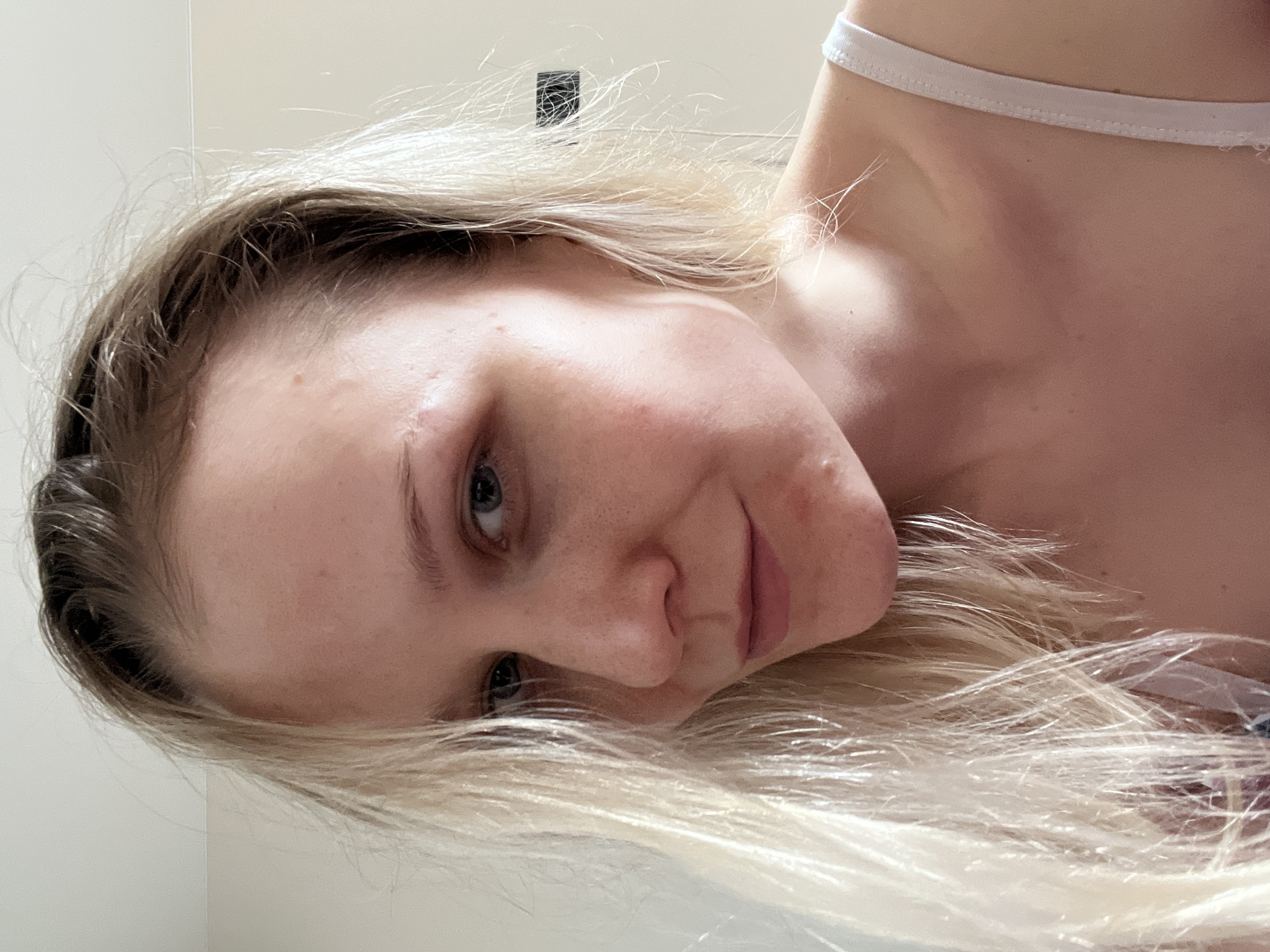 A close-up, angled portrait shows a young woman with light blonde hair. Her eyes are light blue, and her skin appears fair with some minor skin imperfections visible. She is wearing a white strap, suggesting she is in casual attire. The background is a plain, light-colored wall. The image does not reveal any information about the location of the city of Khimki, Russia.