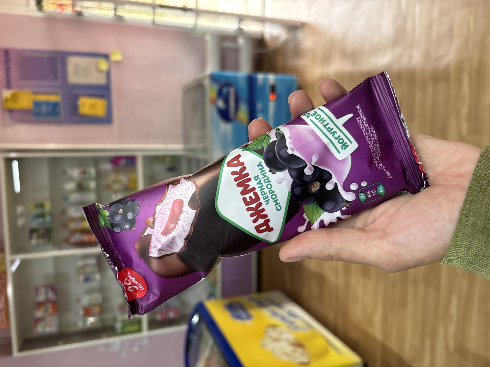 A person's hand holds a purple ice cream wrapper with a picture of blackcurrants and a depiction of the ice cream inside. The wrapper is labeled in Russian with the brand name "ДЖЕМКА" (Dzhemka) and "ЧЕРНАЯ СМОРОДИНА" (Blackcurrant). Below this, the words "ЙОГУРТНОЕ" (Yogurt) are visible. Nutritional information is present on the wrapper, showing "57г", "145 ккал", and "6%". The background suggests an indoor retail environment, possibly a small shop or convenience store, with shelves stocked with various products and refrigeration units. The flooring is wooden. The image is likely taken in the city of Nevon, Russia, given the Russian text on the product.