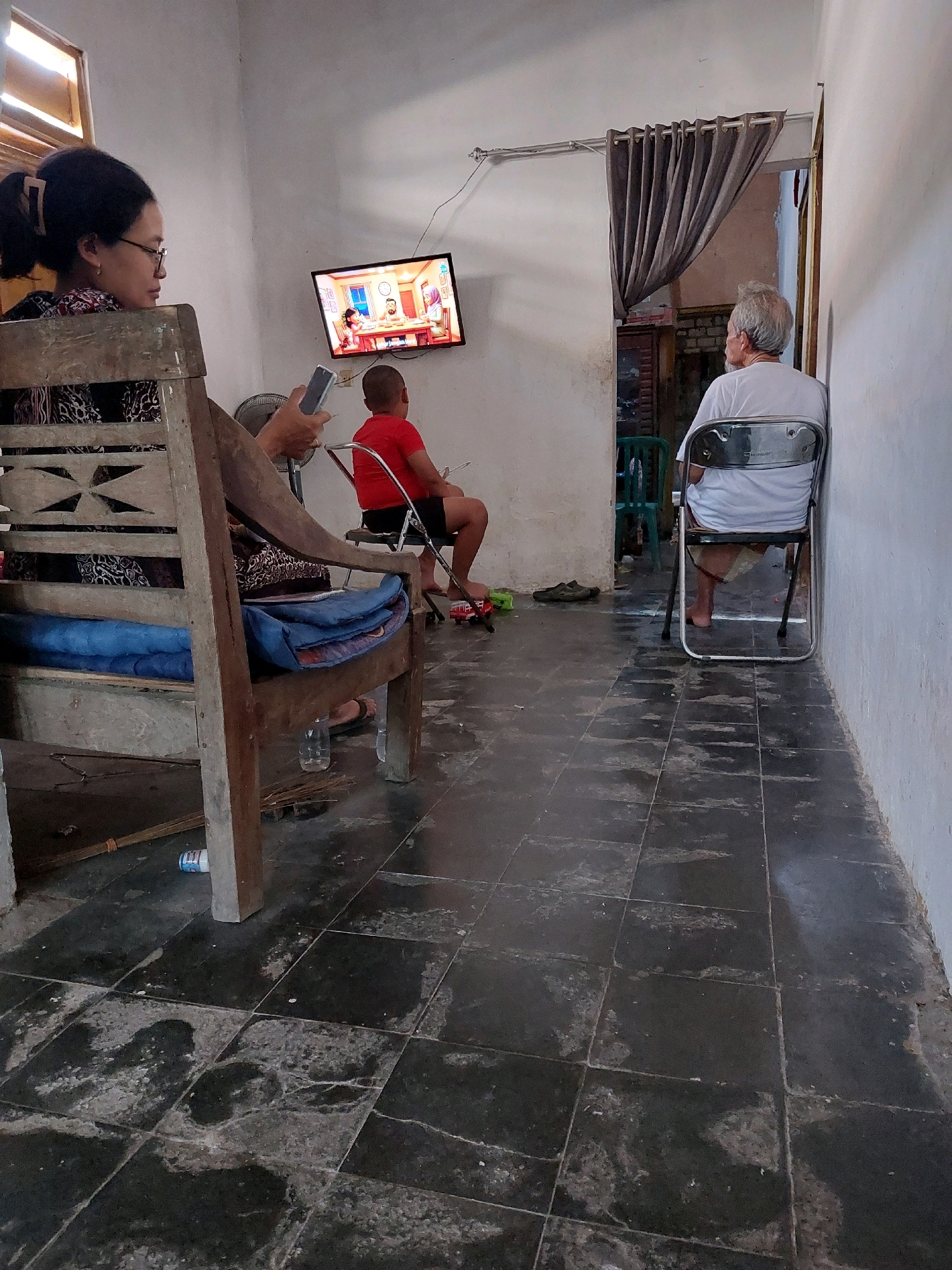 In a sparsely furnished room in Sarangan, Indonesia, three individuals are present. A woman, seated on the left, is looking at a mobile phone she holds in her right hand. She is wearing glasses and has her hair tied back. Next to her, a young boy in a red t-shirt and black shorts sits on a folding chair, facing a television screen mounted on the wall. The television displays a cartoon or animated program. On the right, an elderly man with gray hair is seated on another folding chair, facing away from the viewer, towards an interior doorway where a gray curtain hangs. The floor is tiled with large, dark gray tiles, some of which appear worn or damaged. A wooden bench with a blue cushion is positioned to the left of the frame, and a broom lies on the floor in the foreground. The walls are painted a light color.