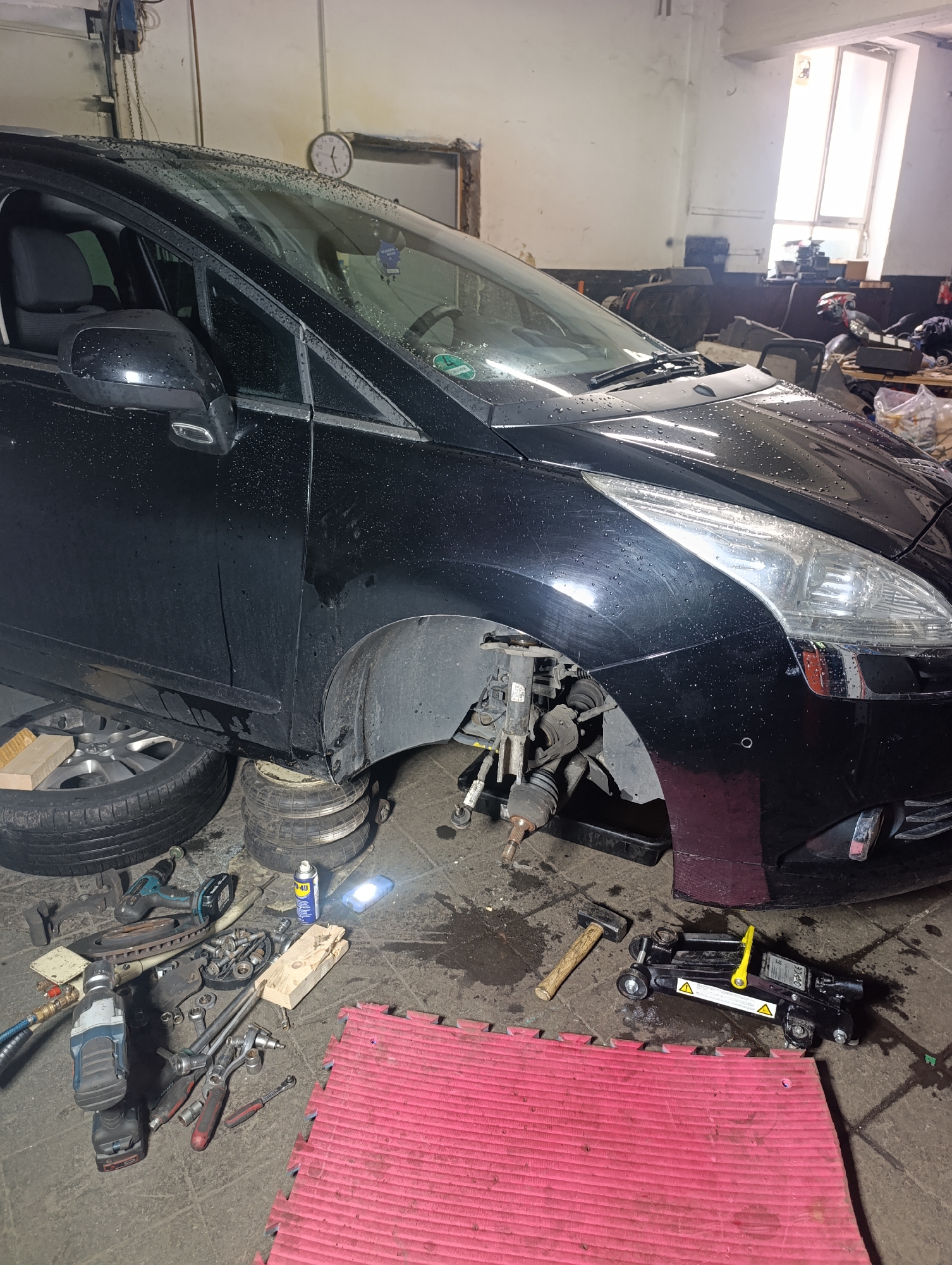 An interior view of a workshop or garage shows a black car, possibly a van, with its front passenger-side wheel removed and the suspension components exposed. The vehicle is supported by stacked inflatable jacks and a wooden block. Tools, including a cordless drill, wrenches, a hammer, and brake rotors, are scattered on the concrete floor around the car. A can of WD-40 and a red mat are also visible. The workshop environment appears somewhat cluttered, with a window in the background that lets in daylight. This scene is indicative of automotive repair work.