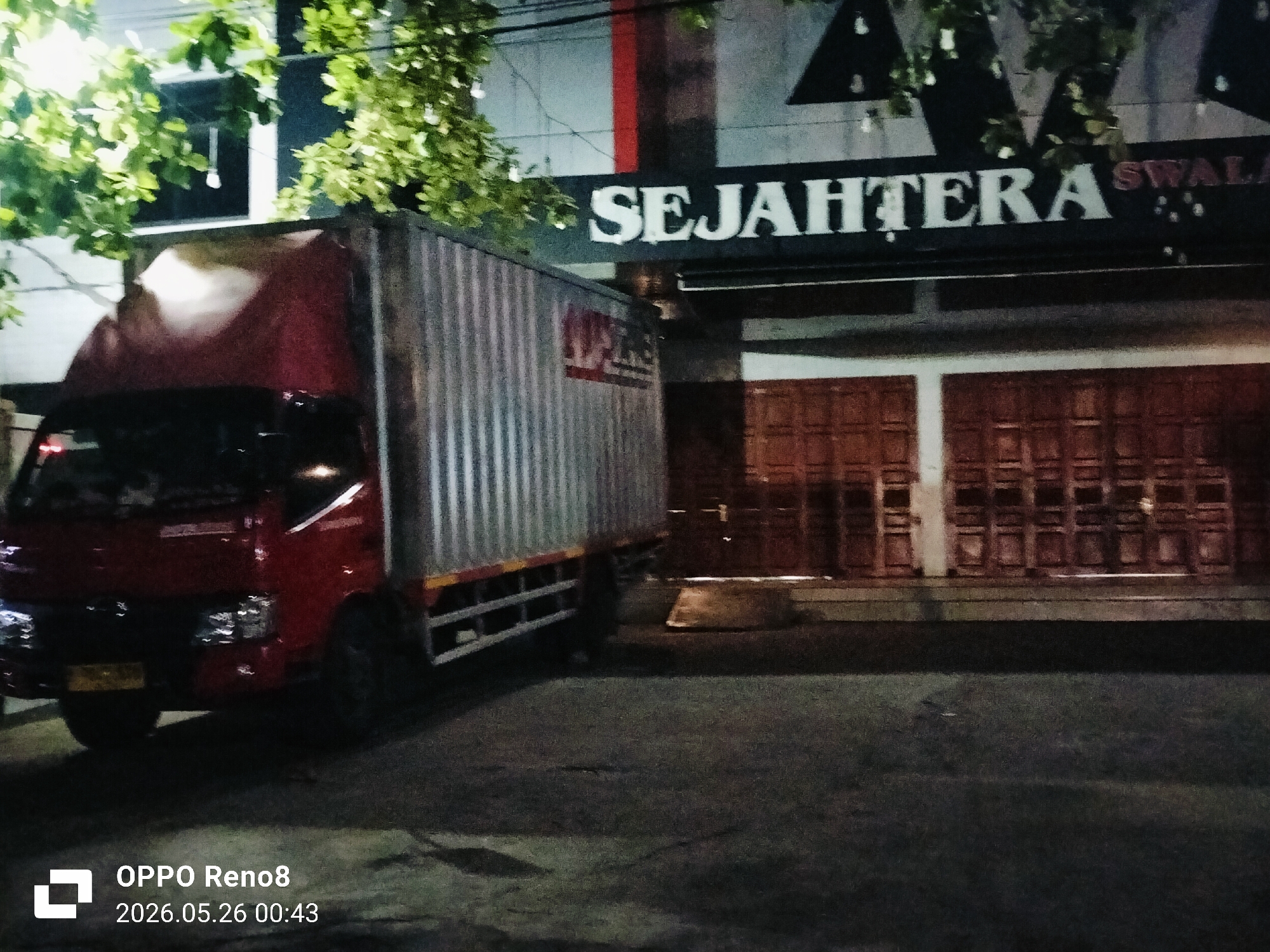 At night in Lhokseumawe, Indonesia, a red and silver delivery truck is parked on a paved surface in front of a building with a sign that reads "SEJAHTERA SWALA". The building has dark wooden shutters or doors. Green foliage hangs over the scene from above. The timestamp "OPPO Reno8 2026.05.26 00:43" is visible in the lower-left corner.