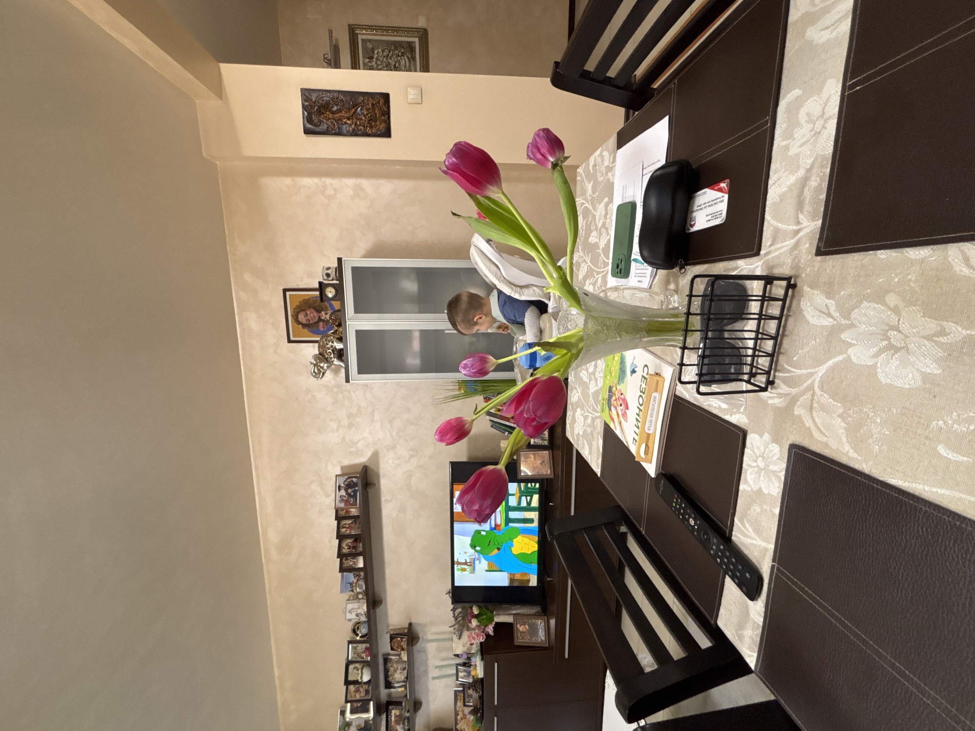 A medium shot, indoors, depicts a dining table scene with a young child seated in the background, likely in Plovdiv, Bulgaria. The table is covered with a patterned tablecloth and several dark brown placemats. Dominating the foreground is a vase of tulips, alongside various objects including a book written in Cyrillic script ("СЕЗОНИТЕ" is visible), a smartphone, a black glasses case, a small black wire rack, and a remote control. In the background, a television displays a cartoon, positioned above a cabinet, part of a shelving unit displaying framed photos. The presence of Cyrillic writing on the book and the general domestic setting suggest the location is likely in Bulgaria. The child in the background is in a highchair and appears to be interacting with something in front of them.