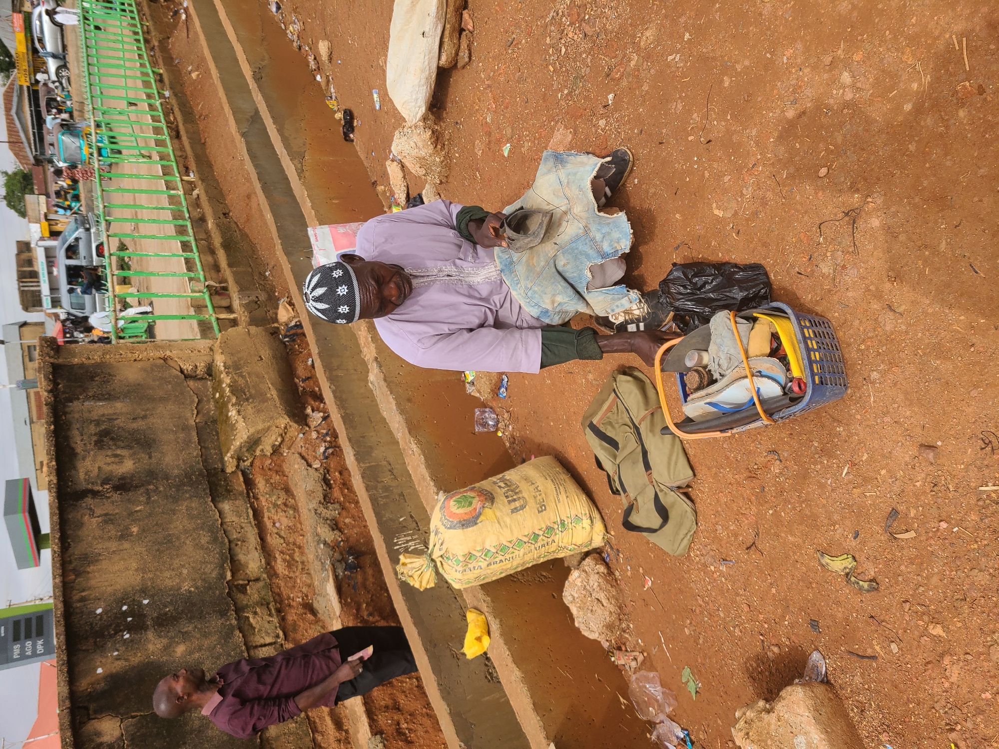A street scene, likely in Unknown, Unknown, shows an older man wearing a white prayer cap and light purple robe sitting on the ground near a concrete curb and green metal guardrail. He is beside a blue plastic crate containing various items. Next to him is a black plastic bag, a beige canvas bag, and a large yellow sack labeled "UREA." A man in a dark maroon shirt walks past the sack, looking at his phone. Debris and litter are scattered across the reddish-brown ground. A gas station sign is visible in the background.