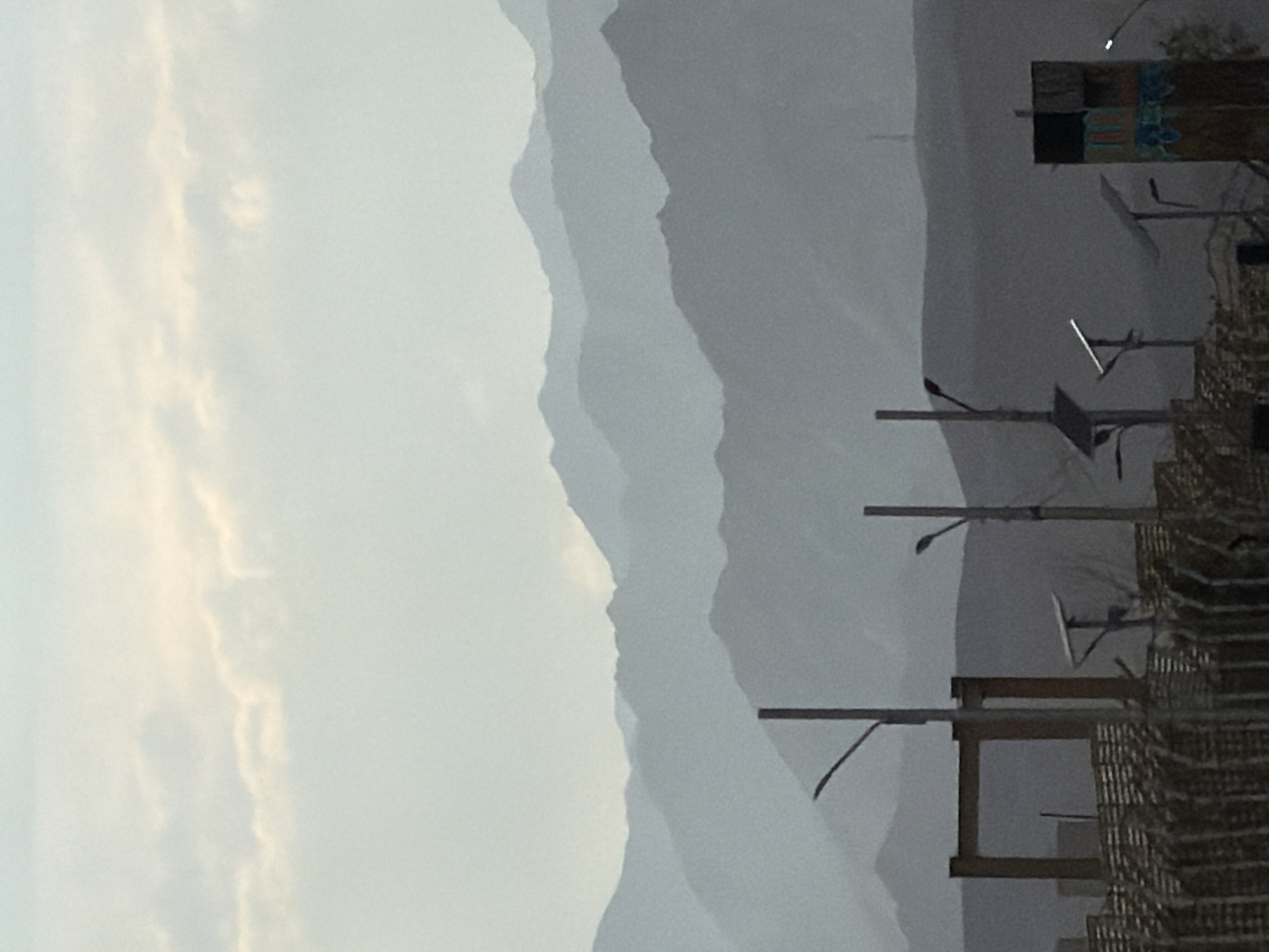 The image shows a hazy, mountainous landscape, likely in the city of Chumkar, India. In the foreground, there are several man-made structures, including a fence, a large rectangular frame, and streetlights. There are also solar panels visible. The mountains in the background are layered and faded due to the atmospheric haze. The sky is overcast with some lighter patches of clouds. There are no people visible in the image.