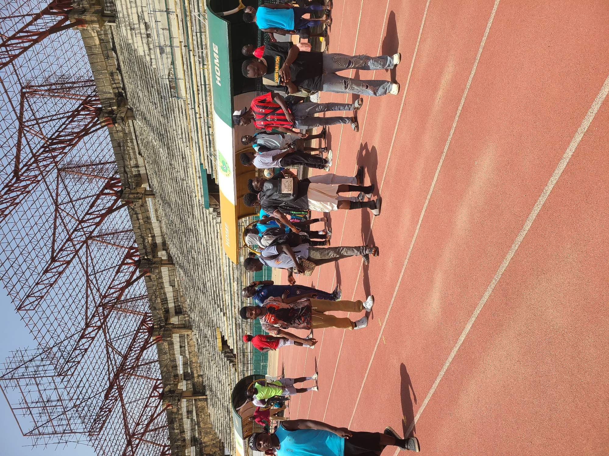 A group of approximately 13 people stand on a red running track adjacent to stadium seating under a partially covered area labeled "HOME". The stadium appears old, with bare concrete seating and rusted metal infrastructure visible on the left. Most of the people in the image are wearing casual clothing; specific apparel noted include a red and black striped shirt, a black shirt with "Louis Vuitton" printed on it, and a blue tank top. The setting is possibly the New Stadium in Nigeria, based on the prompt. The overall scene appears to be outside on a sunny day, as evidenced by the shadows cast on the track.