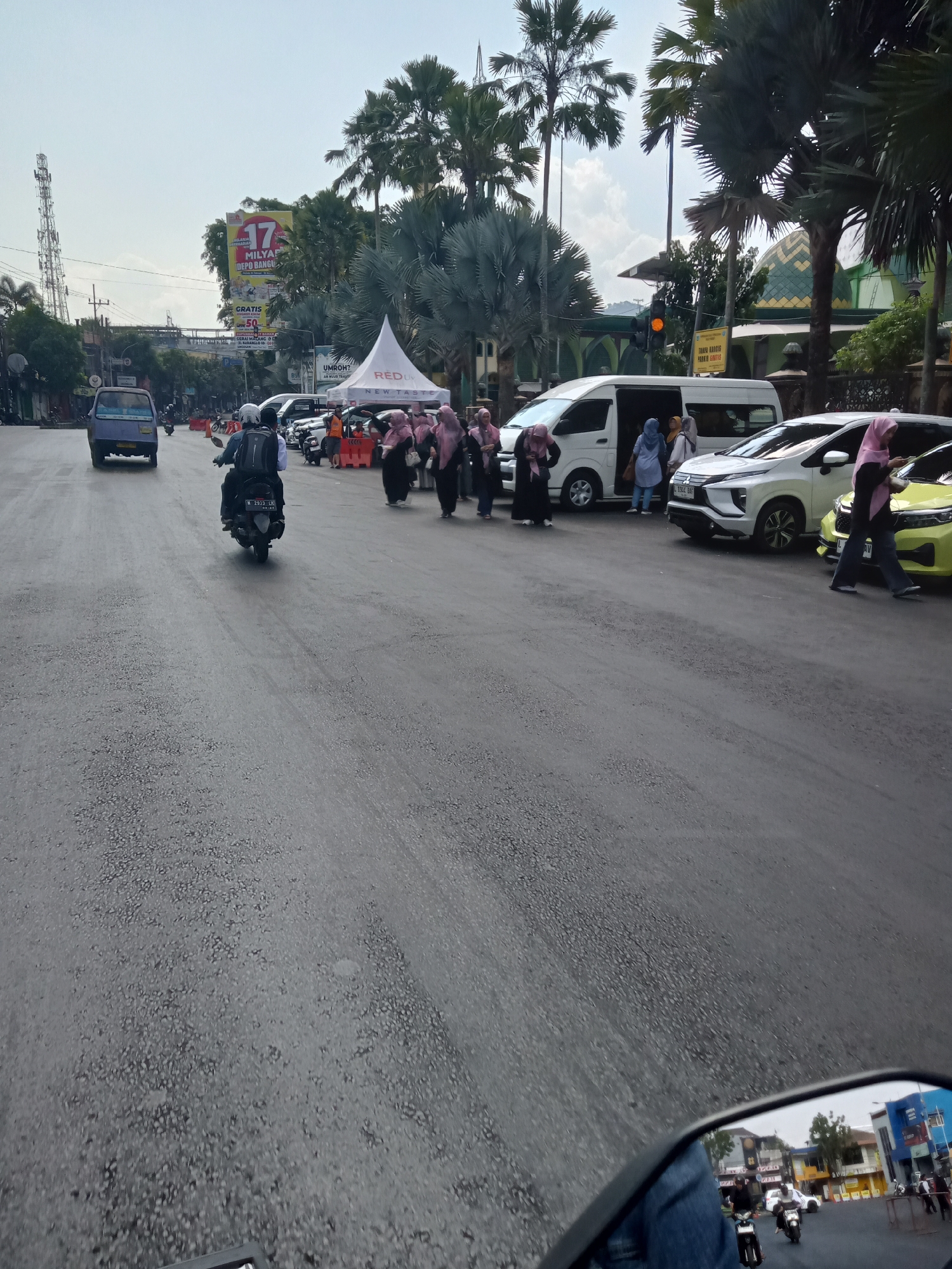 The image depicts a street scene in Batu, Indonesia, with a focus on vehicles and pedestrians. In the foreground, a motorcycle is seen from behind, with its rider wearing a helmet and a backpack. The road surface appears wet and reflective, suggesting recent rain. To the right of the motorcycle, several cars are parked or moving slowly, including a white van with its doors open and people disembarking. A group of women wearing headscarves and dark clothing are walking on the sidewalk. Further down the street, a purple van is in motion. In the background, a large billboard with Indonesian text and graphics is visible, along with palm trees and the dome of a building that resembles a mosque. The sky is overcast. The scene appears to be a busy urban area, possibly near a commercial or event space, given the presence of tents and the billboard.