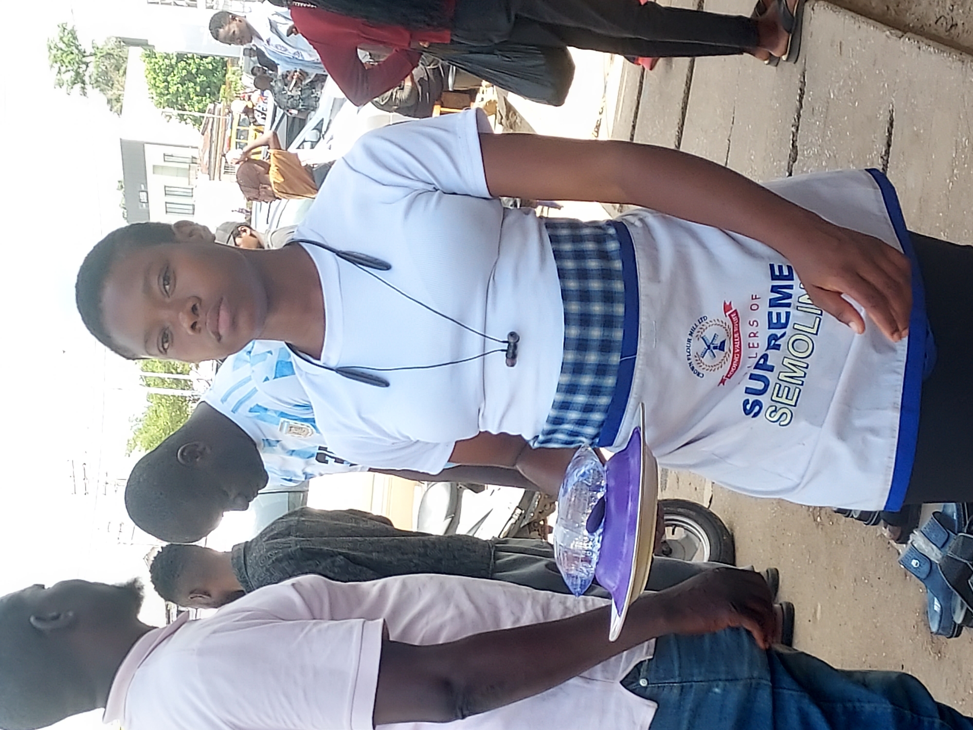 The image captures an outdoor scene, likely a busy street or public area in Jos, Nigeria, during daylight hours. The central figure is a young woman, seen from the waist up, facing the camera. She has short, dark hair and a neutral expression, wearing a white short-sleeved t-shirt and an apron. The apron is white with a blue plaid pattern on the upper part, and a blue trim along the bottom edge. Wired earbuds hang around her neck.
On the apron, prominent text reads "SUPREME SEMOLINA" in blue block letters. Above this, a circular logo contains smaller text, including "CROWN FLOUR MILL LTD" and "STRONG VALUE".
Behind her, several other individuals are visible. To her left, a man in a light pink polo shirt and jeans is partially seen, holding a purple and clear tray-like object. Further back, another man in a light blue and white striped jersey (possibly an Argentina football kit) is looking down. Other people, some obscured or partially visible, are in the background, suggesting a bustling environment.
Elements of the setting include the paved ground, a building with windows in the distance, and some green foliage from trees. A part of a vehicle, possibly a motorcycle or scooter wheel, is visible near the bottom right. The overall impression is that of a public, informal gathering or promotional activity, given the woman's branded apron.