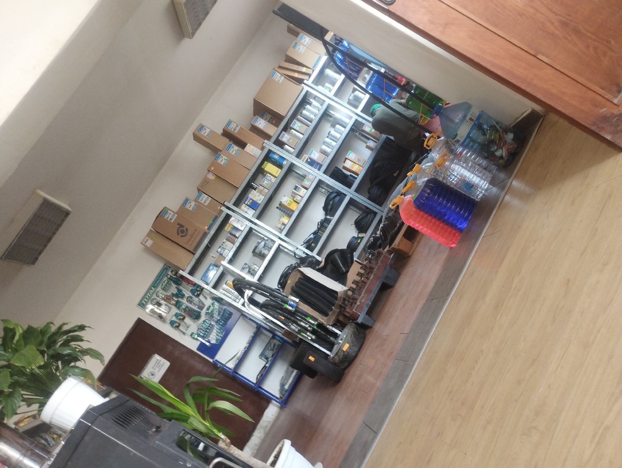 The image depicts an indoor scene, possibly a storage area or a retail space, with a distinct tilt to the right. In the foreground, the polished wooden floor transitions to a greyish-blue floor at the edge of the room. Several large plastic bottles, some filled with water and others with what appear to be colorful objects, are arranged on the floor. Two of these bottles are red and blue, stacked on top of each other.
The midground features rows of metal shelving units filled with various items. The shelves on the right are packed with cardboard boxes, many labeled, and some smaller packages or containers. The shelves on the left display a wider variety of items, including what appear to be tools or hardware on a display rack, black bags, and more boxes. A tall shelving unit with blue accents holds numerous items, some of which are clearly visible as bottles and containers. In the middle of the room, black hoses or cables are bundled together and leaning against a metal frame. Two tires are also visible in this area, one with an orange tag.
The background includes a dark object that could be a piece of furniture or equipment, next to a potted green plant. A brown door is visible on the right side of the image. The walls are painted in a light, neutral color, and the ceiling has lighting fixtures. The overall impression is one of a functional space where goods are stored or displayed, possibly a workshop, a warehouse, or a specialized store. The scene is likely located in the city of Sredets, Bulgaria, given the context. There is no clear indication of specific actions or interactions between people, though a person in dark clothing is partially visible in the background, crouching near the shelves.