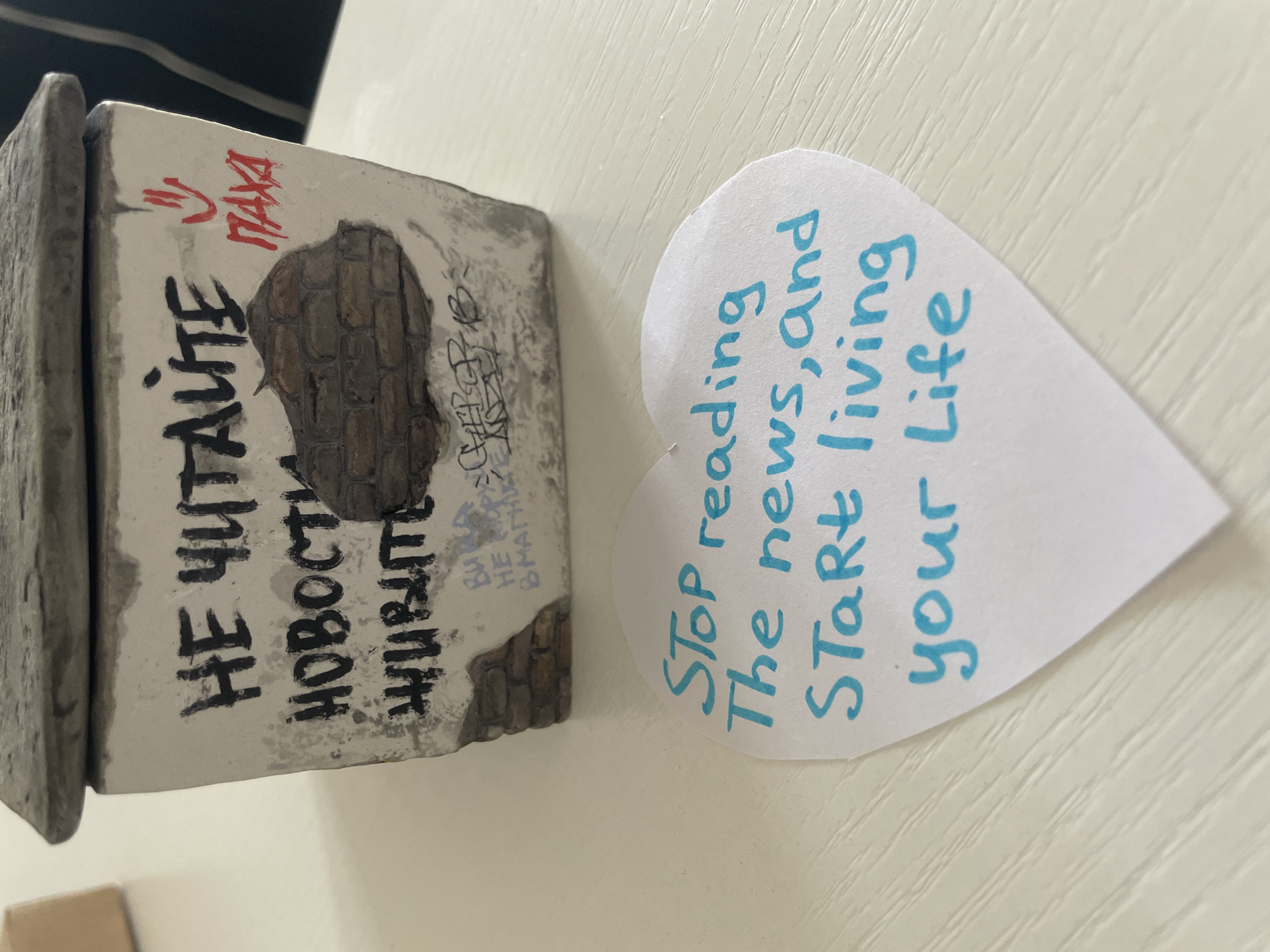 The image shows a small, rectangular, cement-like object with faux brick detailing and graffiti-like markings, next to a heart-shaped piece of paper.
The cement object is textured with an imitation brick pattern and graffiti that includes:
* "НЕ ЧИТАЙТЕ" (Don't Read)
* "НОВОСТИ" (News)
* "ЖИВИТЕ" (Live)
* "ВЫ НЕ В МАГНИТ" (You Are Not Magnet)
* A signature-like mark with "ΠΑΧΑ" and a smiley face above it.
The heart-shaped paper has the following text written in blue ink: "Stop reading the news, and start living your life."
Based on the content of the written words in Russian and English, it appears these items are set indoors, sitting on top of a light coloured surface, possibly a table or desk.