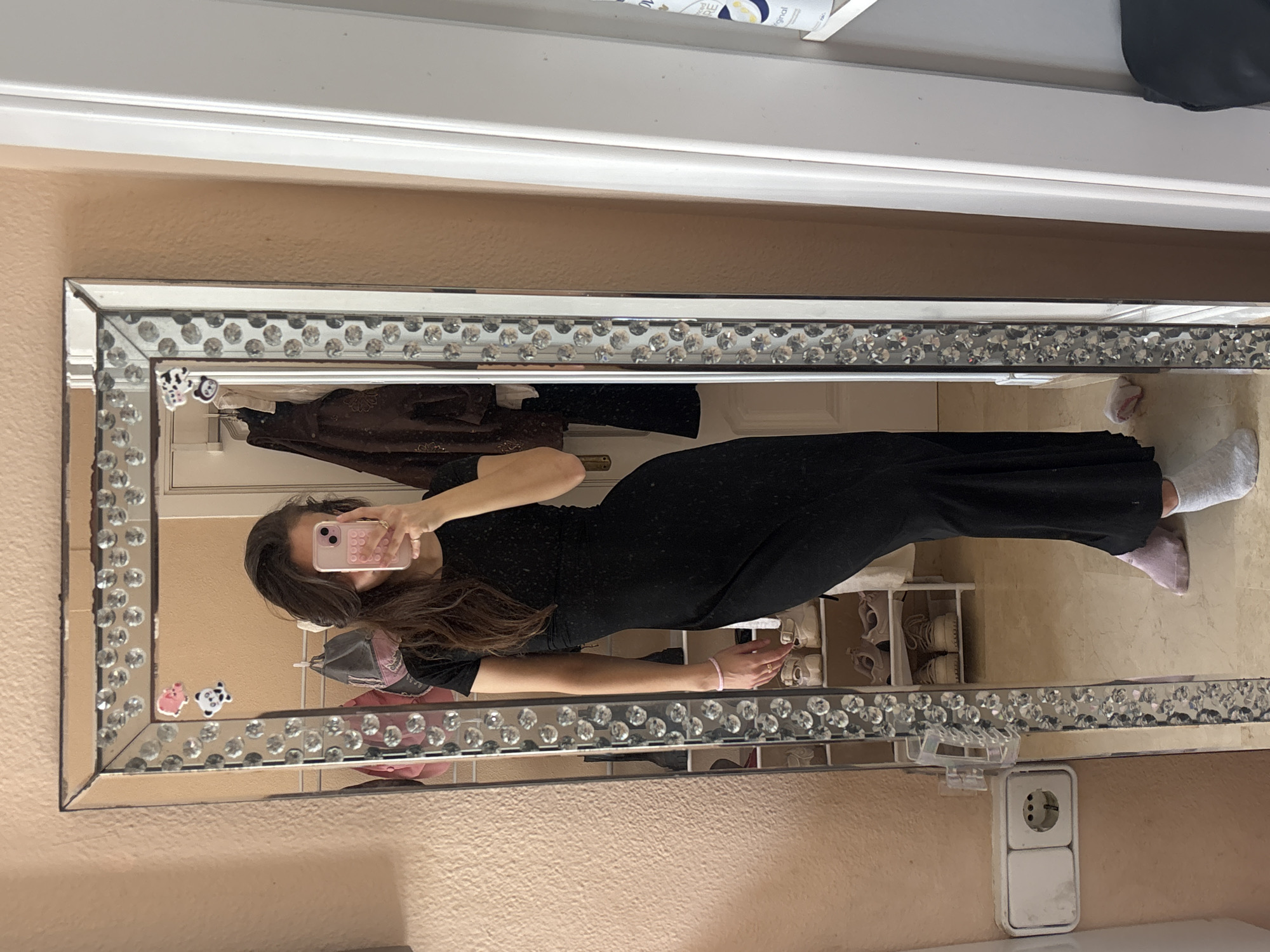 A young woman stands in front of a decorative mirror, holding a pink cell phone in her right hand, taking a selfie. She is wearing a black, floor-length dress with short sleeves. Her hair is long and dark brown, and it falls over her shoulders. She is wearing two socks, one pink and one grey, and has a pink bracelet on her left wrist. The mirror is tall and rectangular, with a frame decorated with clear rhinestones. A pink pig sticker and a black and white panda sticker are attached to the top left of the mirror frame. In the reflection of the mirror, a shoe rack with several pairs of shoes is visible, as are clothes hanging on a rack. A light peach-colored wall is visible behind the woman and the mirror. To the left of the mirror, a white electrical outlet with two sockets is mounted on the wall. To the right of the mirror, a white door frame and a white shelf with a white bottle on it are visible. The floor appears to be tiled. The scene is indoors, likely in a bedroom or hallway. There is no information about the location in Orihuela, Spain.