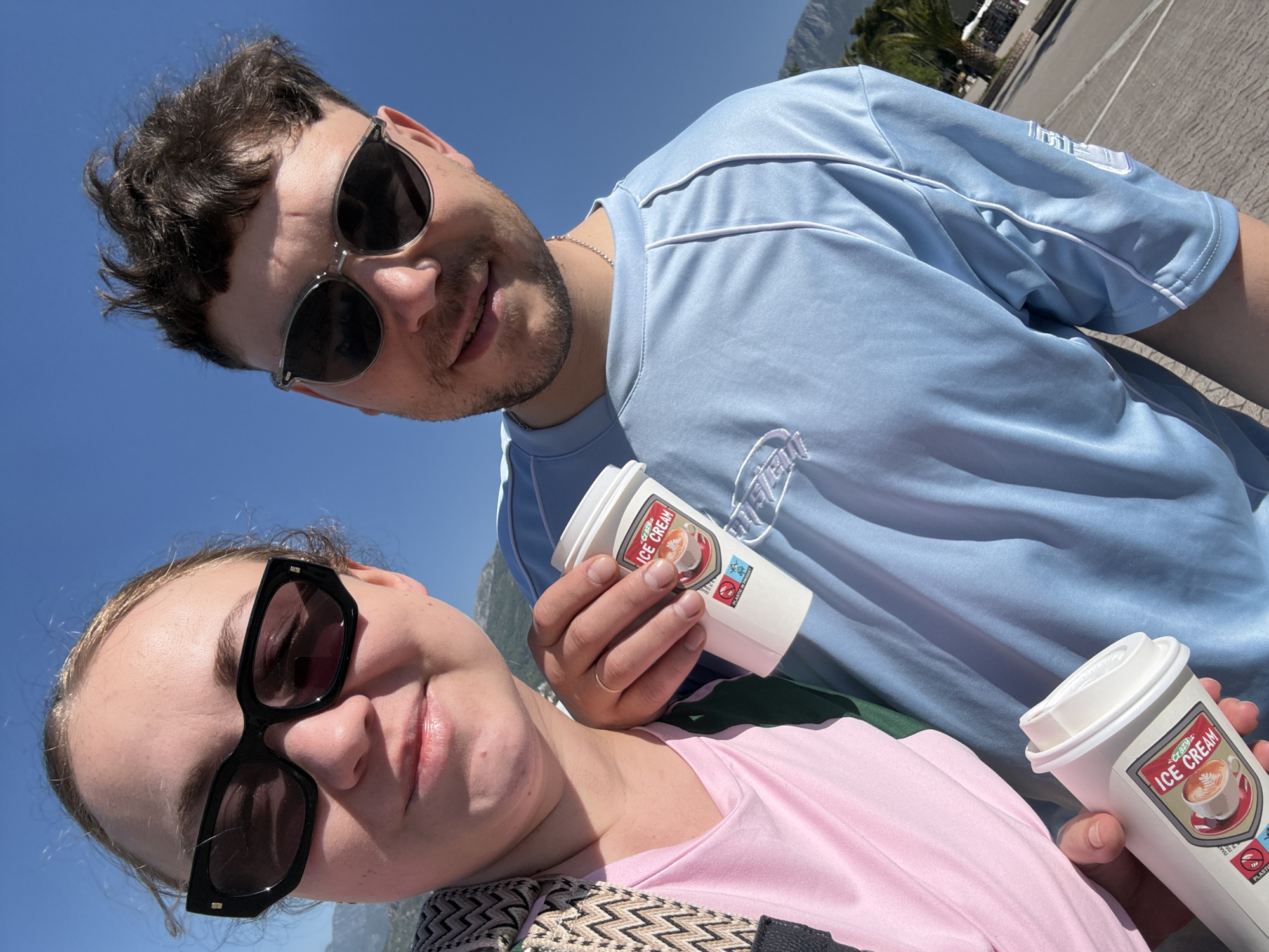 A low-angle medium shot captures two people holding ice cream cups. The man, positioned slightly above and to the right, wears black sunglasses, a blue shirt with white piping, and a silver chain. He holds a white ice cream cup in his right hand with a logo that reads "Crazy Ice Cream." The woman, angled below and to the left, wears black sunglasses and a pink shirt. She is holding another ice cream cup with a lid. A shoulder strap is visible across her body. The background includes a clear blue sky, a mountain range, and some greenery suggesting a sunny outdoor setting. The location is likely Bar, Montenegro.