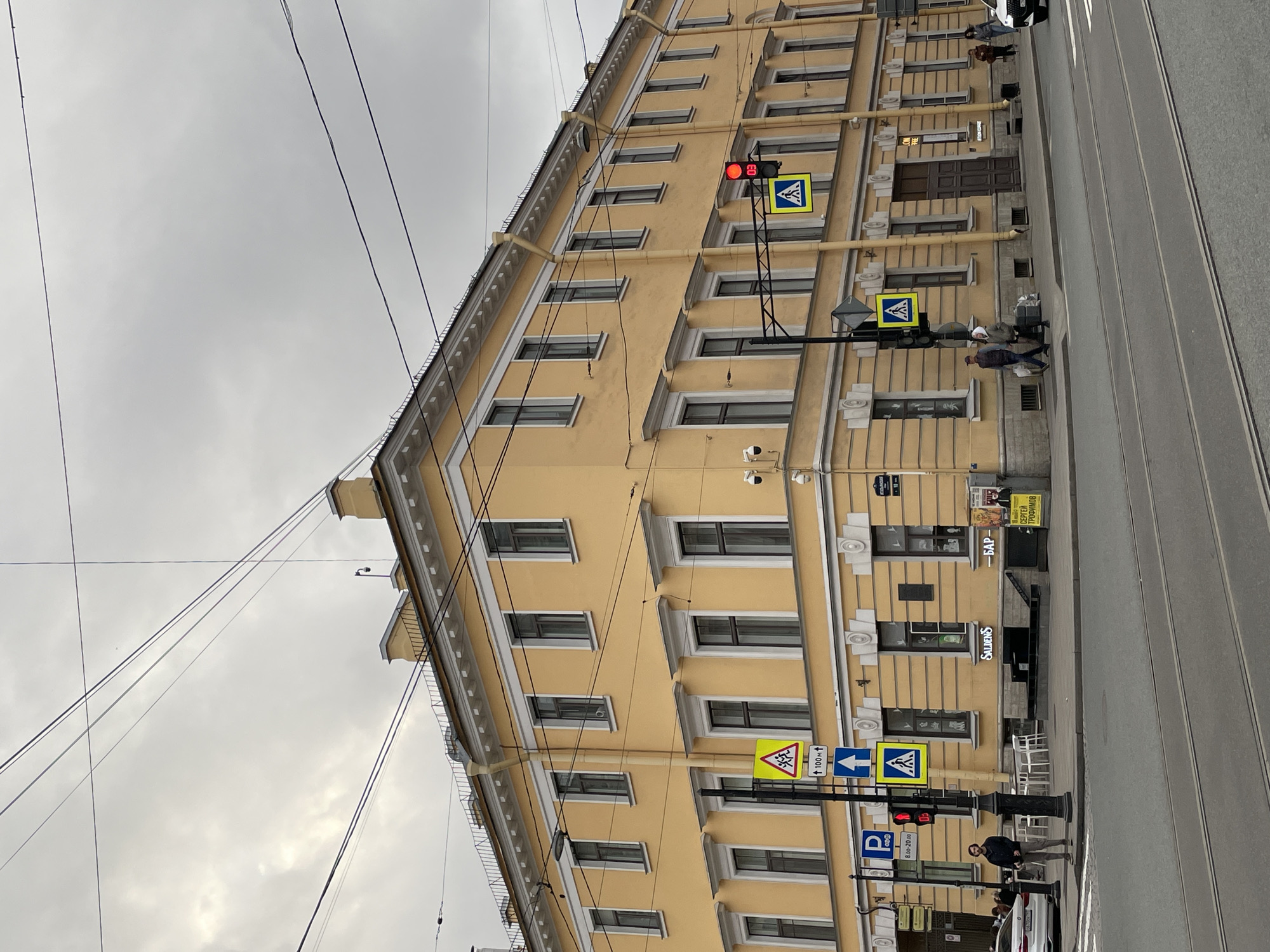 A street scene in Saint Petersburg, Russia, shows a tall, pale yellow building with multiple windows and a decorative cornice. Traffic lights are red, and pedestrian crossing signs are visible. Power lines run across the top of the frame, against a gray, overcast sky. People are walking along the sidewalk. Street signs and advertisements are posted on the building. A business called "Saldens" is located on the ground floor of the building. A white car is parked at the side of the street. The street appears to be paved with tram tracks.