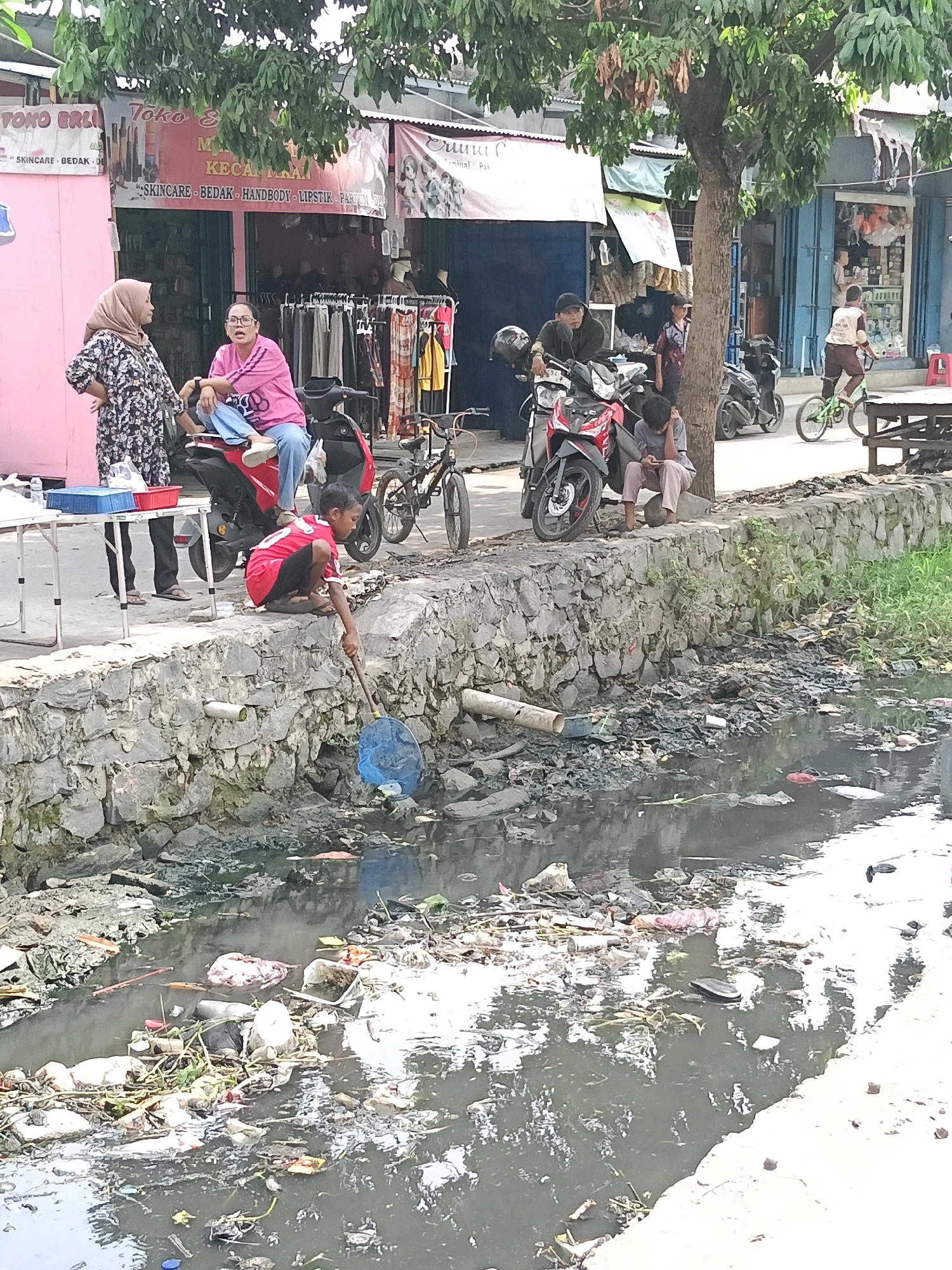 The image depicts a street scene in the city of Belimbing, Indonesia, adjacent to a polluted canal. A stone wall separates the street from the canal, which is filled with a large amount of garbage, including plastic bags, bottles, and other debris.
On the street, several individuals are present. Two women are standing near a red motorcycle, one wearing a hijab and a floral dress, the other in a pink shirt and blue pants. A young boy in a red shirt and black shorts is crouching by the canal wall, holding a blue net, appearing to be fishing or scavenging. Further down the street, a young person sits on a rock near a tree, looking at a mobile phone. Several motorcycles are parked along the street.
In the background, shops are visible with awnings and various goods displayed. A child is seen riding a green bicycle on the sidewalk. The overall environment suggests a busy urban area, contrasted sharply by the heavily polluted waterway.