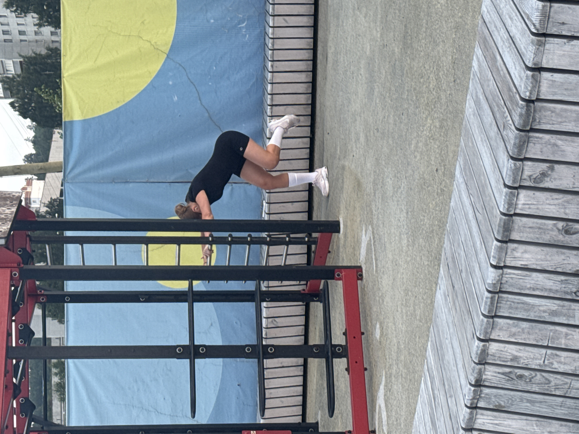 A woman is performing a workout on an outdoor fitness equipment structure. She is wearing a black sports bra and black shorts with white socks and sneakers. She is in a downward-facing dog position with her hands on the ground and her hips raised. The background features a blue and yellow mural and a building. The scene appears to be set during the daytime, possibly on a cloudy day given the lighting. The fitness equipment is black and red metal, with bars and rungs. To the right of the woman is a slatted wooden structure. The overall impression is of an urban outdoor fitness environment. No visible text.