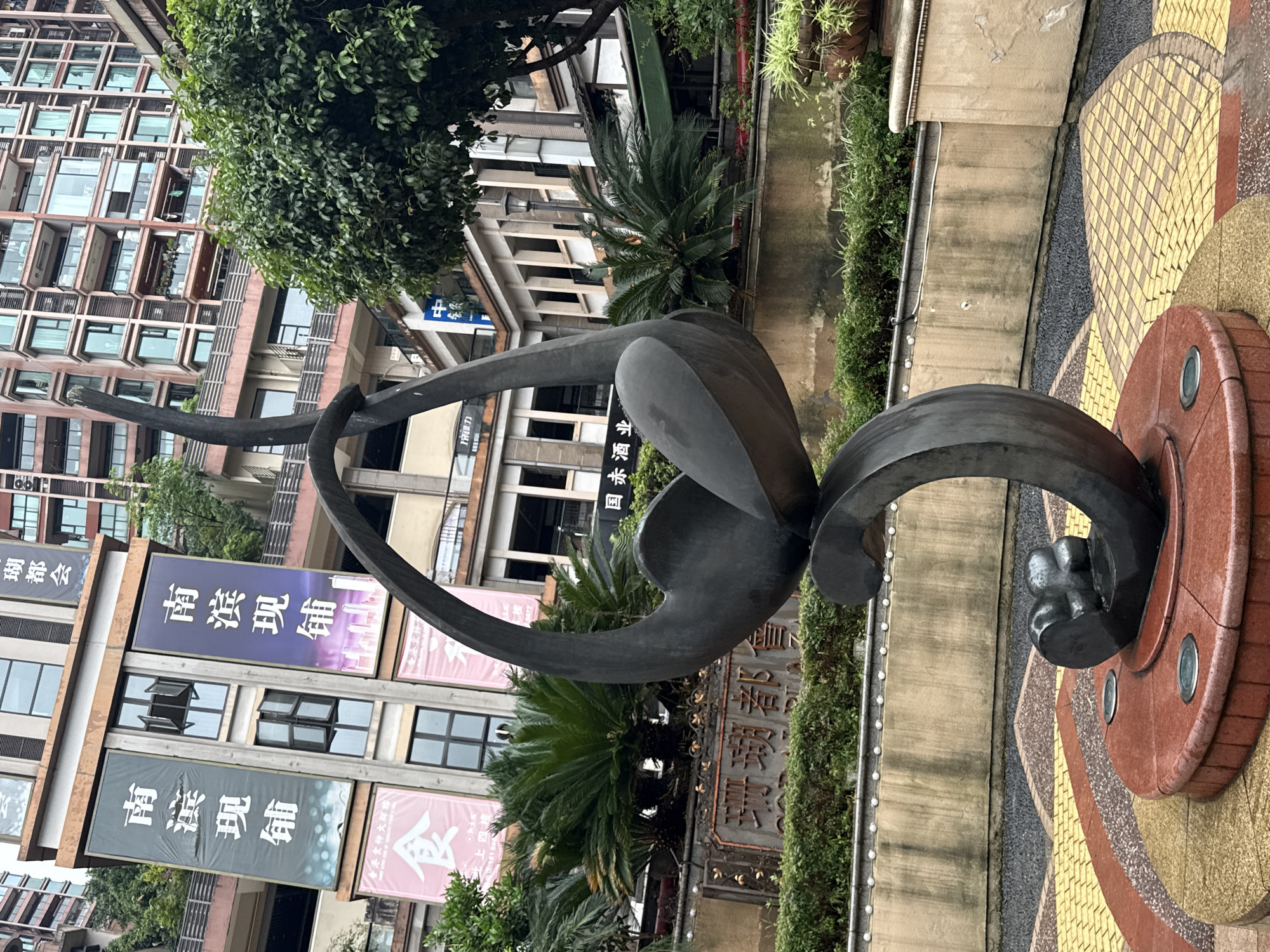 A metal sculpture stands in front of buildings and advertisements in Nan'an District, China. The sculpture is a curving shape, with one section resembling a bird's head or leaf. The base of the sculpture is a circular pattern in shades of brown, yellow, and grey. The buildings in the background are a mix of architectural styles, with banners advertising "Nan Bin Xian Pu" in Chinese characters. Vegetation is interspersed throughout the scene, including trees and potted plants.